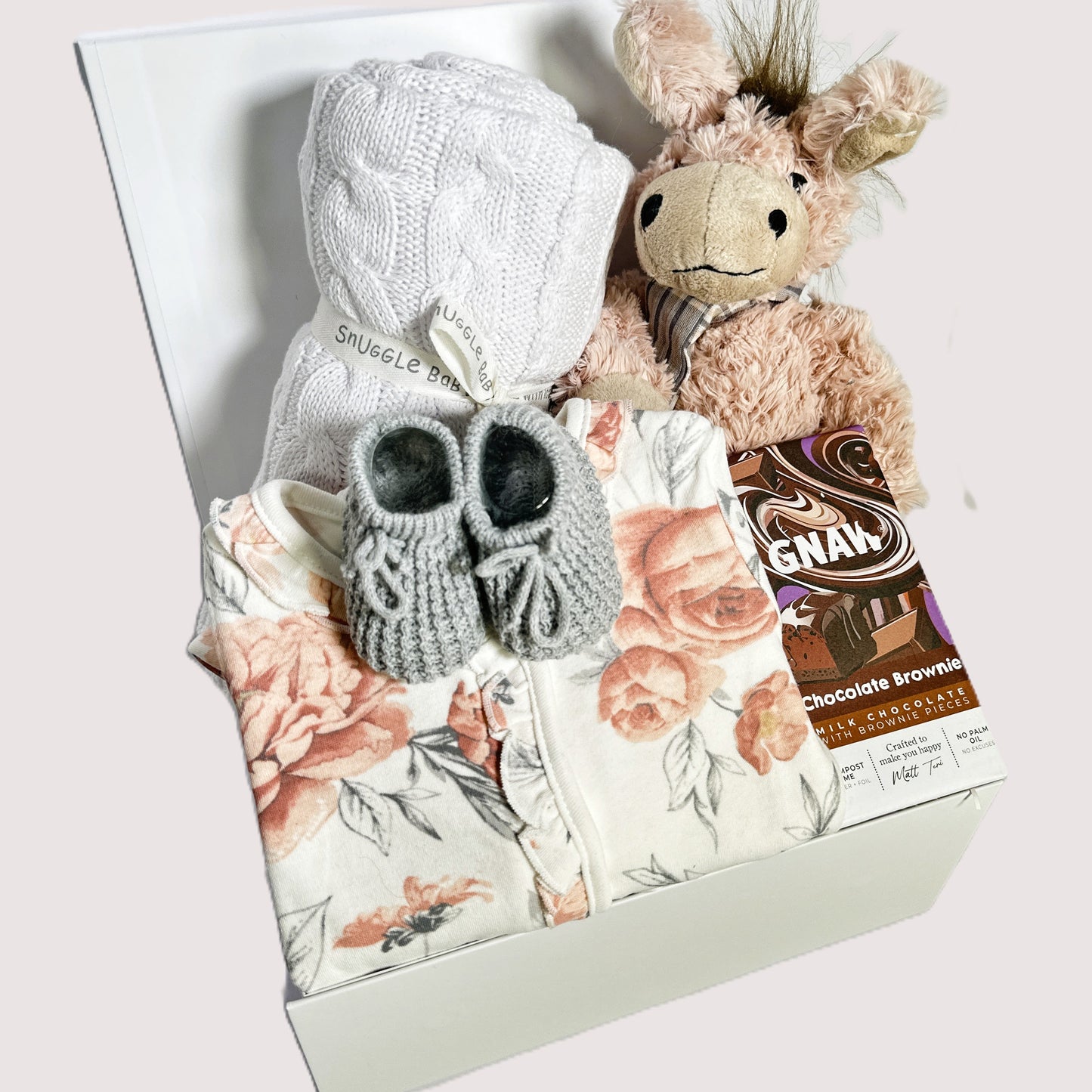 a baby girl hamper gift with a white cotton baby blanket, donkey soft toy, floral zip up sleepsuit, baby booties and a bar of Gnaw chocolate.