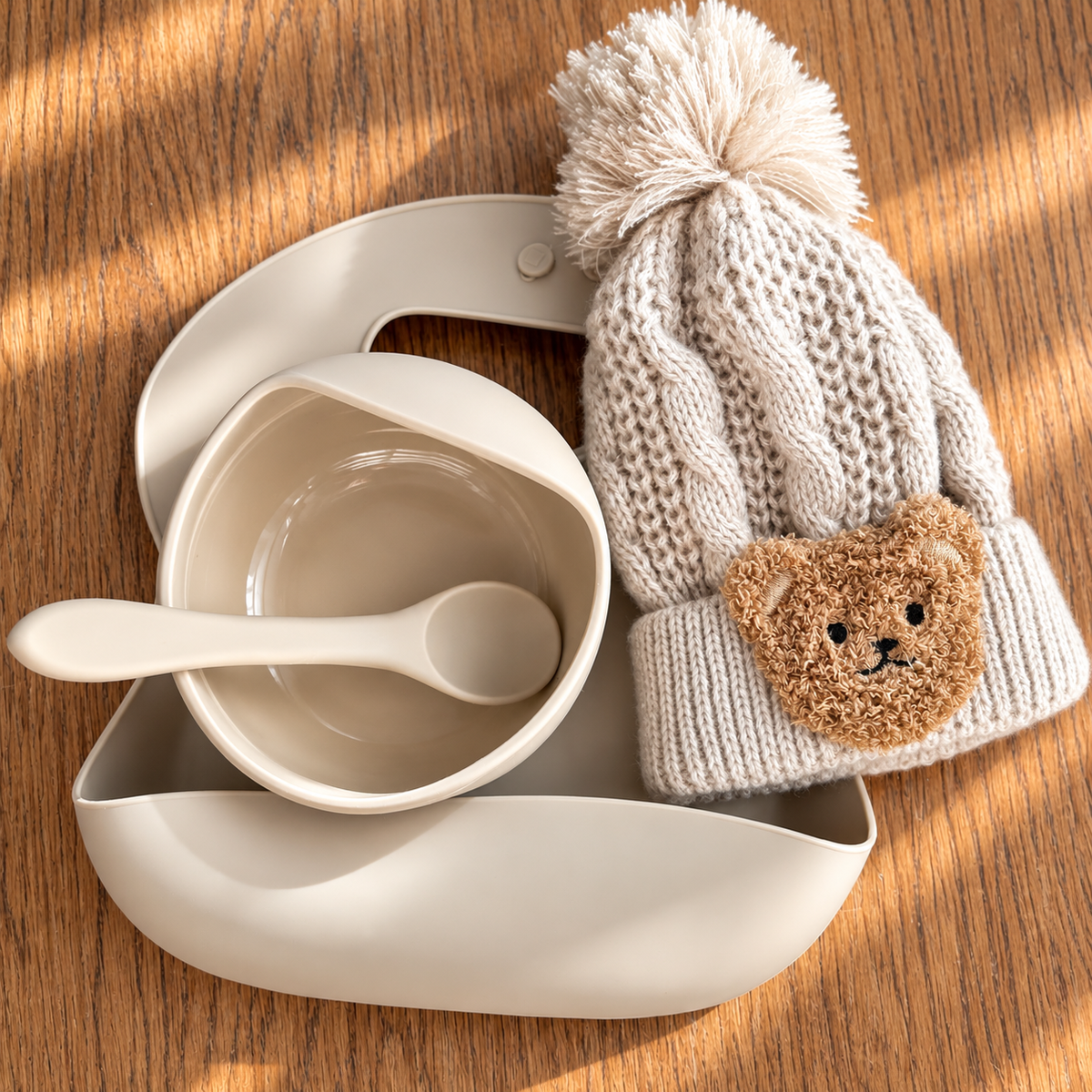 A baby feeding set arranged on a wooden surface, including a cream-colored silicone bowl with a spoon, a matching tray, a knit pom-pom hat, and a teddy bear face applique on the brim.