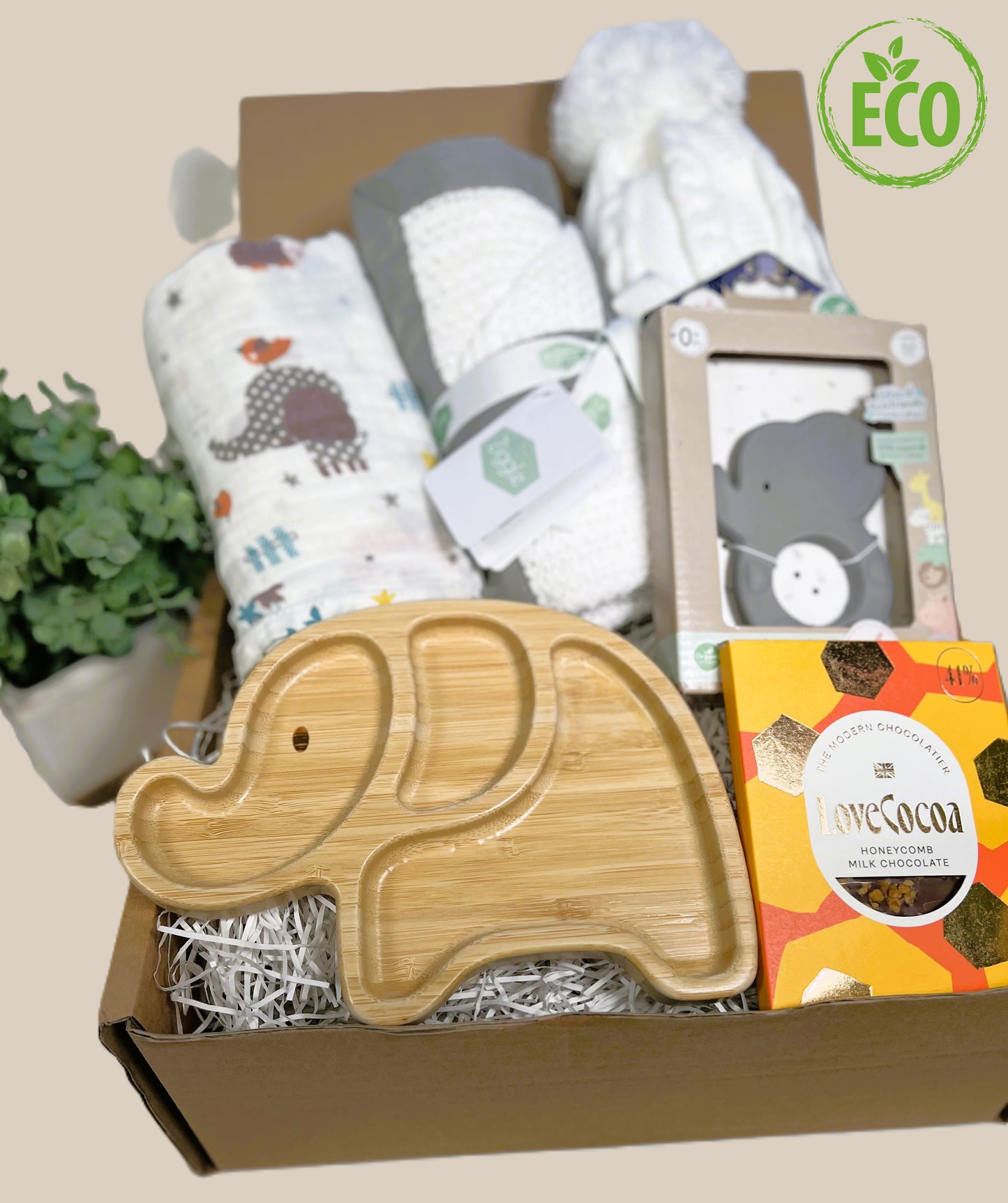 Unisex baby shower gift comprising a bamboo elepant baby plate with silicone suction plate underneath, a 120cm by 120cm muslin swaddling cloth with jungle animal prnt, a white cotton cellular baby blanket with a grey trim, a white baby pompom hat, a Tikiri natural rubb er baby teether in the shape of an elephant and a Love Cocoa bar of honeycomb chocolate