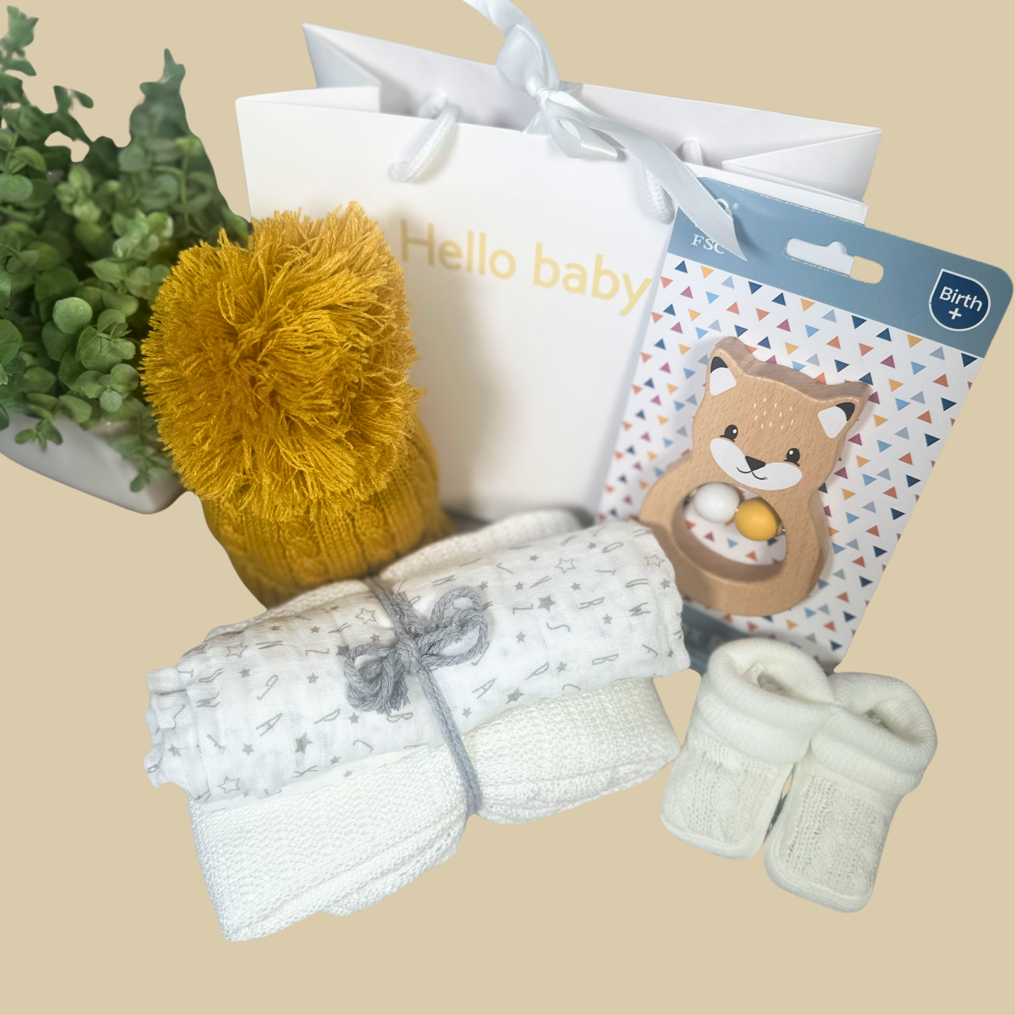 Baby gift hamper featuring a white patterned towel, a pair of white knit booties, a yellow pom-pom baby hat, a white 'Hello baby' gift bag, and a packaged wooden animal teether on a beige background.