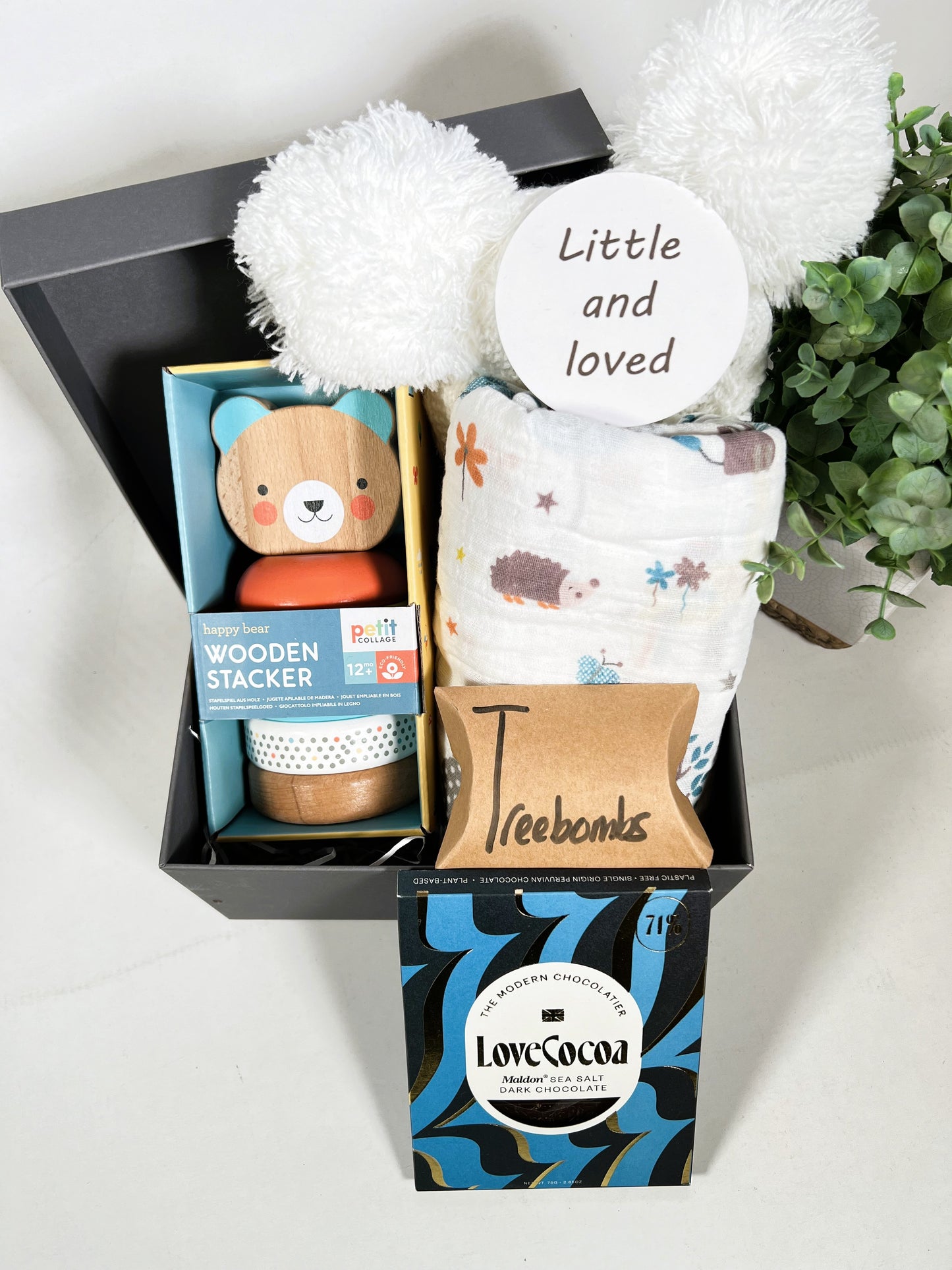 A Lovely new baby gift containing a eco friendly wooden bear stacker toy, a large muslin with jungle print, a white pompom baby hat, a bar of Love Cocoa chocolate, 3 Treebombs and a baby photography disc in a grey square baby keepsake box.
