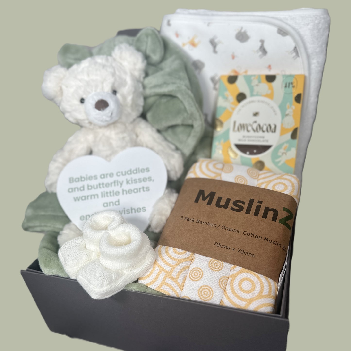 Baby gift set with teddy bear, blanket, and MuslinZ products on a light green background