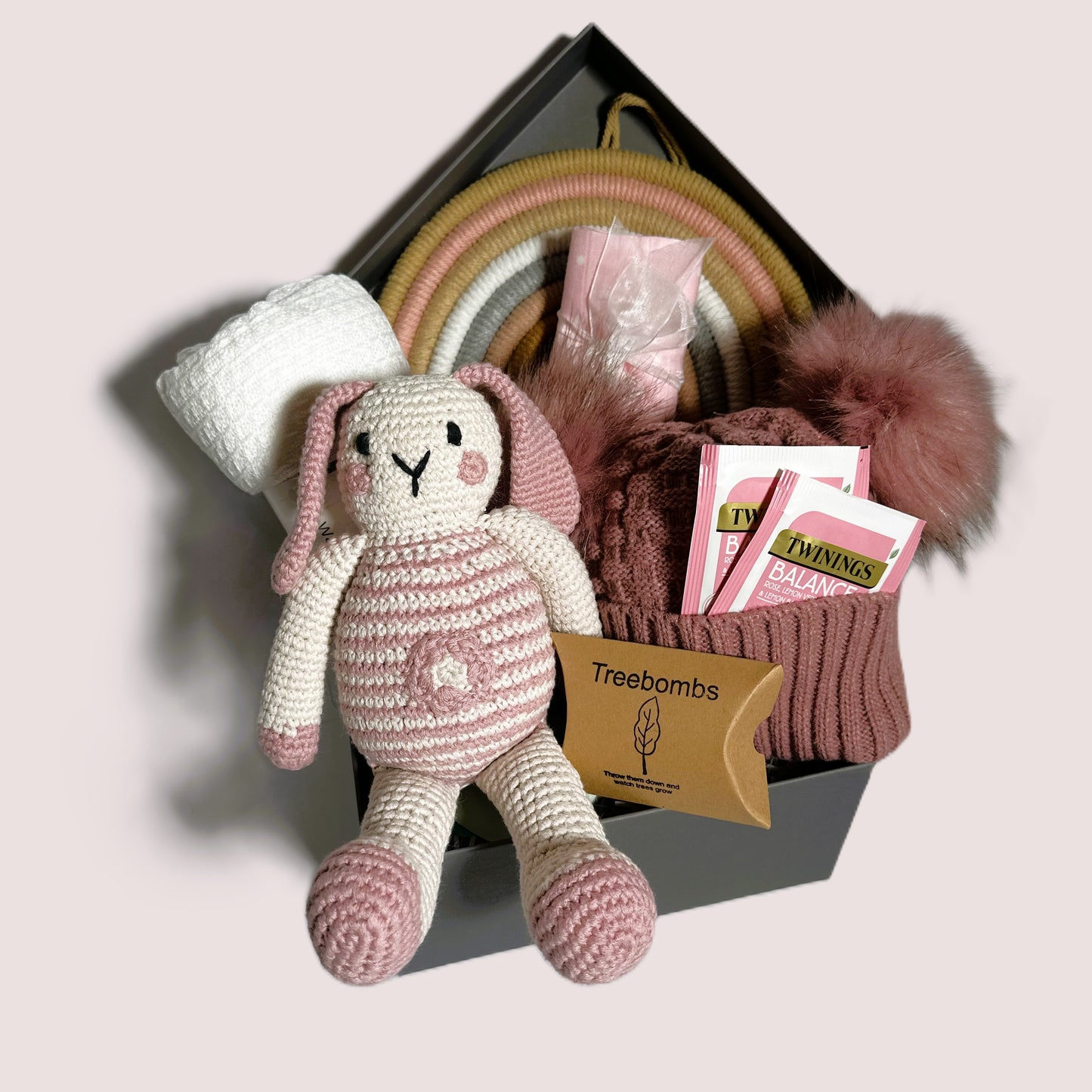 A new baby girl hamper in a grey baby keepsake box containing a crocheted bunny baby soft toy, a white cotton baby blanket, a rose pink baby pompom hat, some Twinings teabags and a pink and white muslin square.