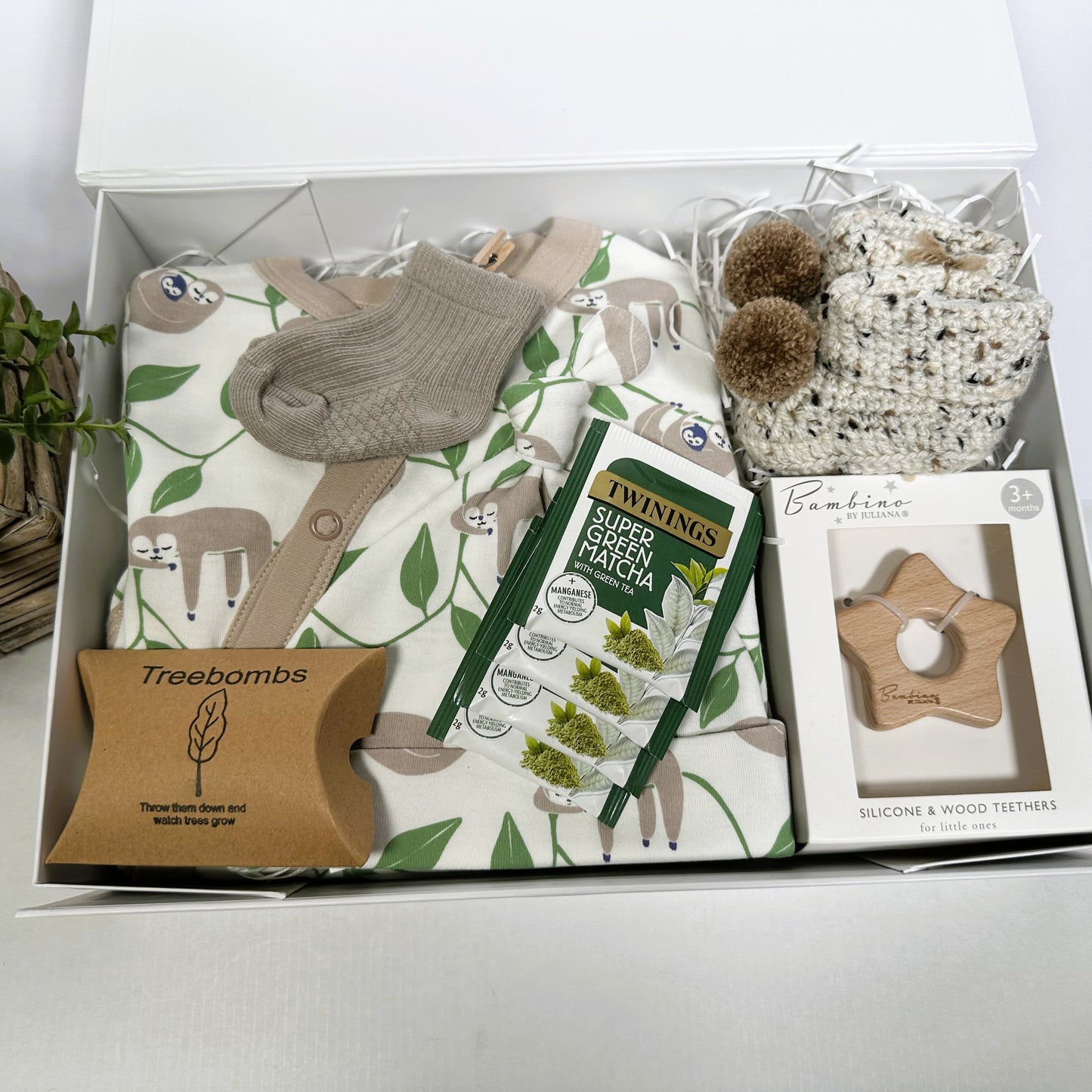 Baby shower gift containing a sloth print baby sleepsuit and baby knot hat, soft, crocheted baby booties, a pair of cotton sock in taupe ,Bambino star teether and some Matcha teabags. All in a white baby keepsake box.