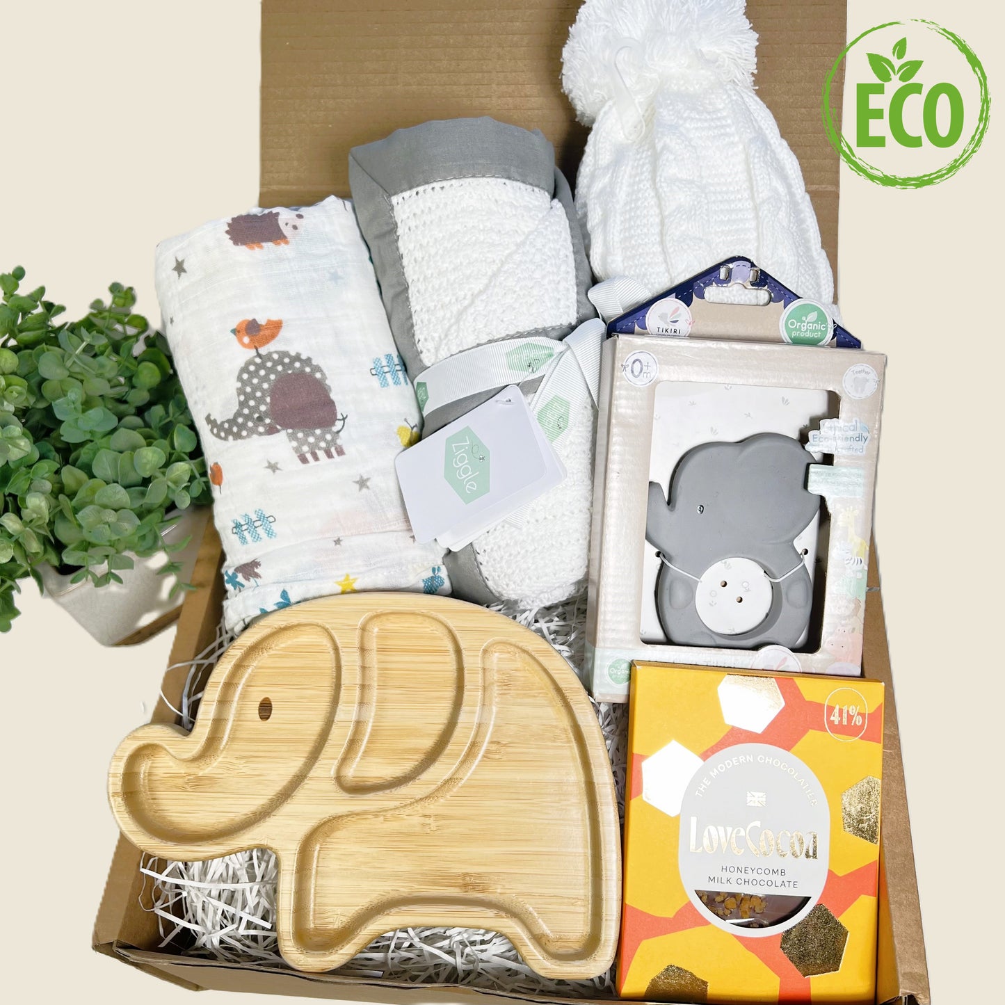 baby shower gift comprising a bamboo elephant baby plate with silicone suction plate underneath, a 120cm by 120cm muslin swaddling cloth with jungle animal prnt, a white cotton cellular baby blanket with a grey trim, a white baby pompom hat, a Tikiri natural rubb er baby teether in the shape of an elephant and a Love Cocoa bar of honeycomb chocolate