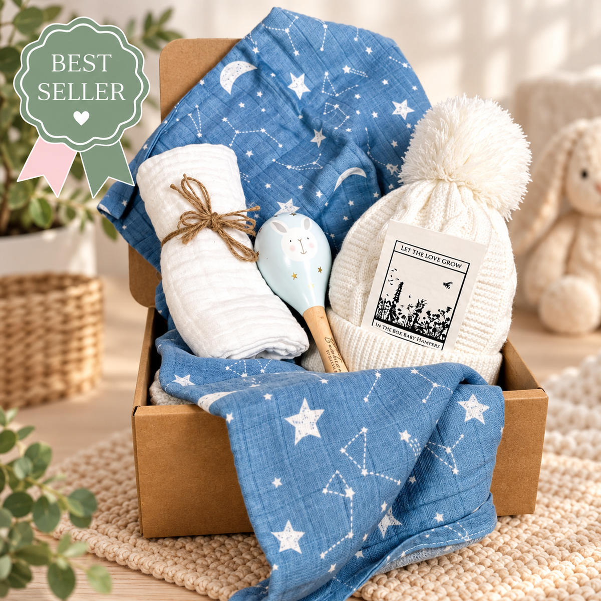 A baby gift set arranged in a brown box, featuring a blue muslin blanket with white star and constellation patterns, a matching white muslin tied with twine, a white knit pom-pom beanie baby hat baby maraca wooden toy. A green 'BEST SELLER' badge appears in the top left, with a soft nursery-style background including a stuffed bunny and plants.