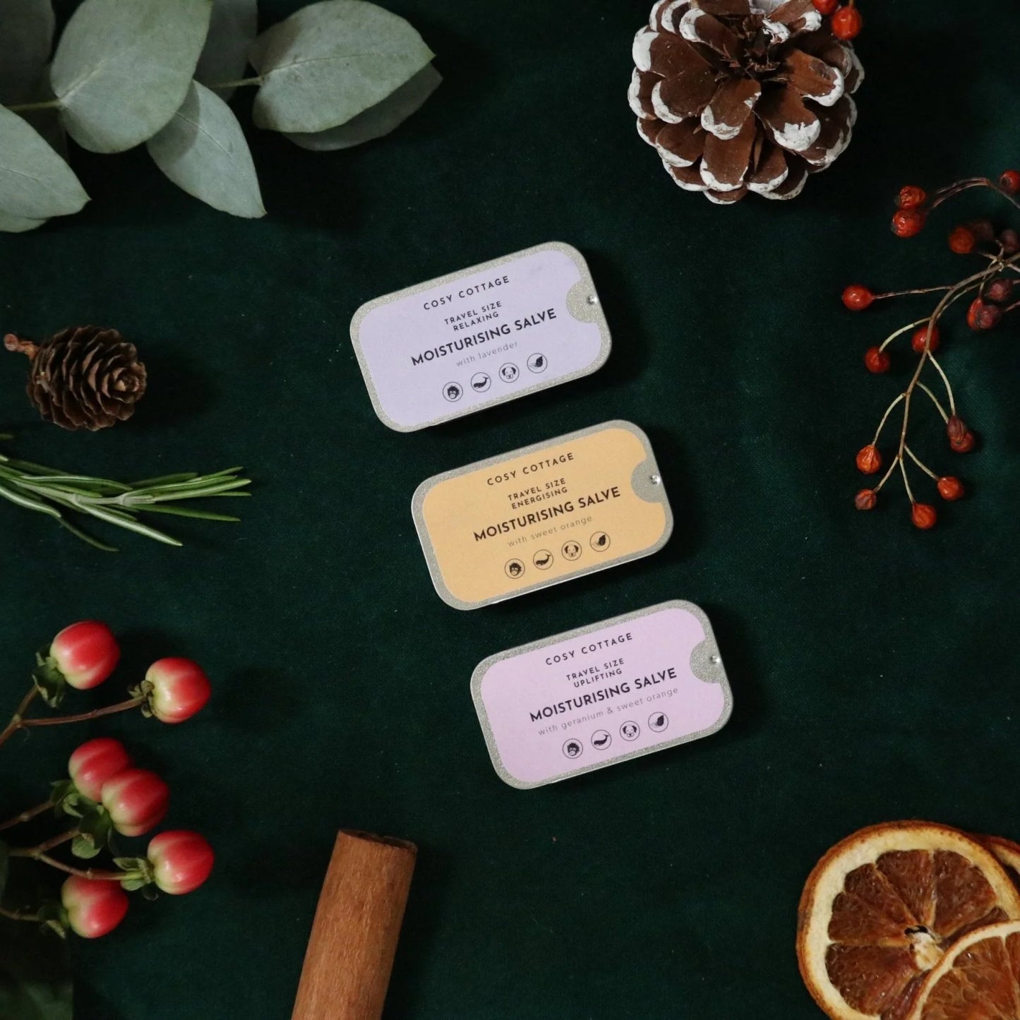Three small containers of Cozy Cottages Moisturising Salve on a dark surface with decorative elements like pine cones and berries.