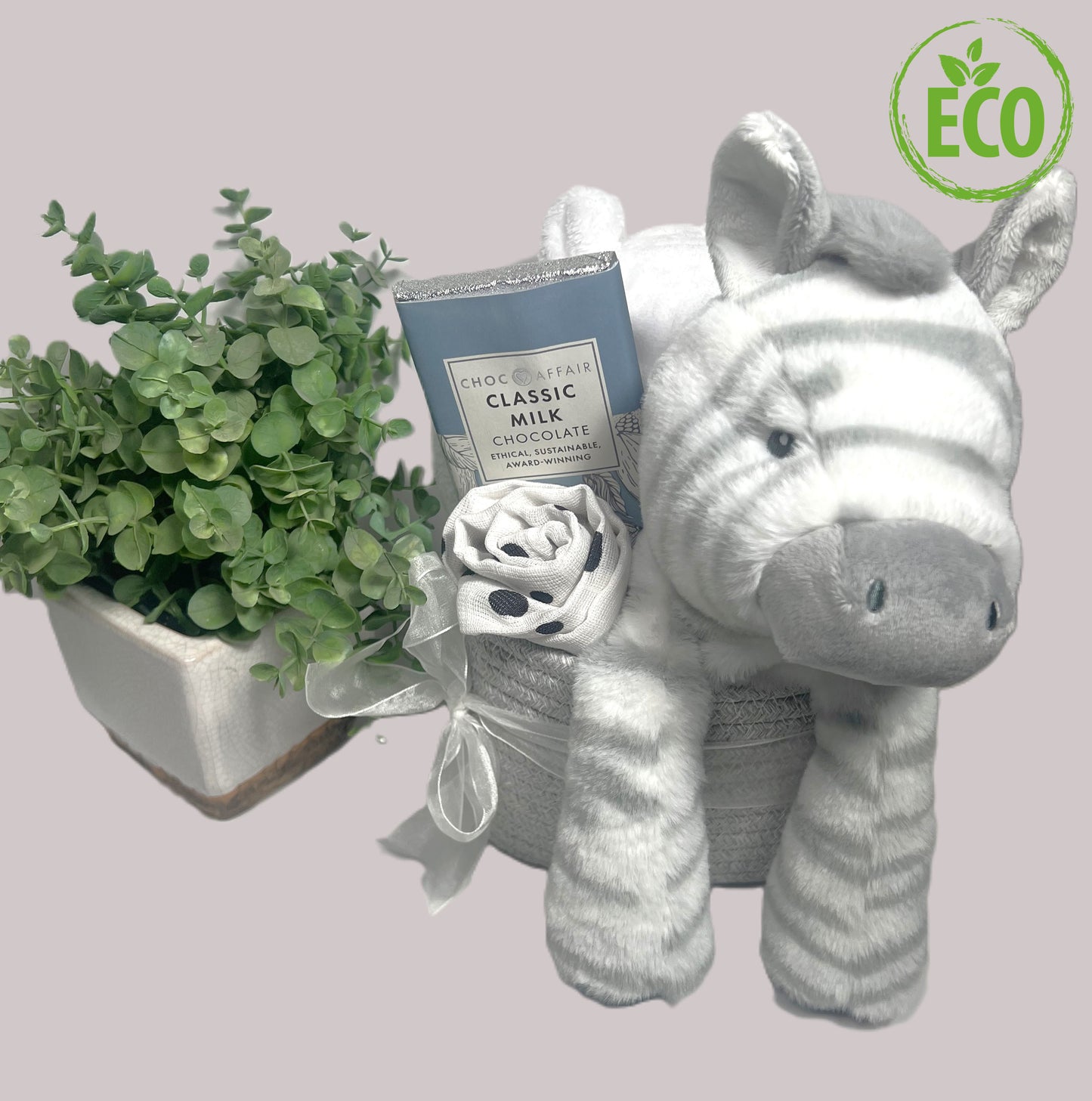 A mummy and baby hamper basket in a rope storage caddy containing a Keeleco 25cm Zebra soft baby toy, a white baby dressing gown , 90gm bar of Choc Affair dark chocolate and a white and black large baby muslin square.