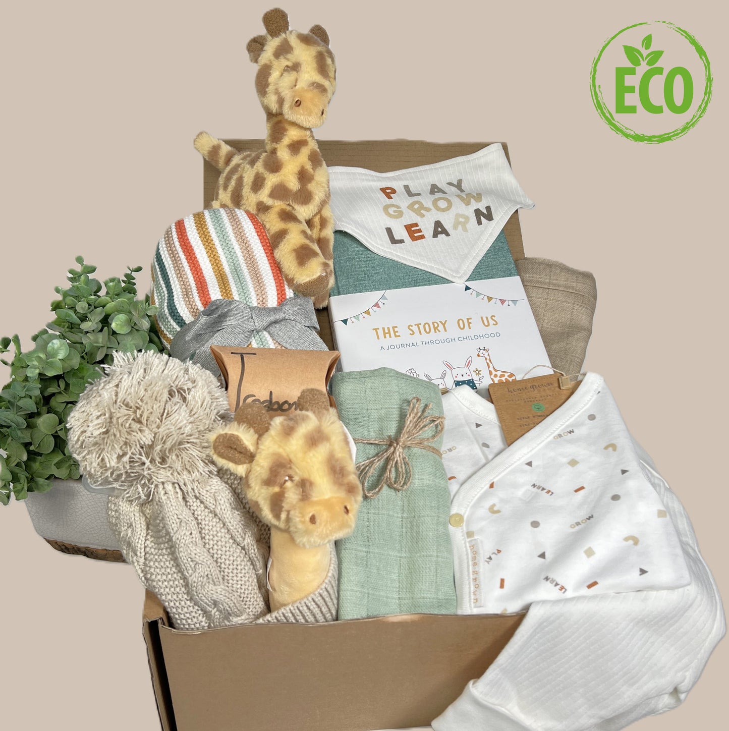 Organic clotton layette set in cream with a neutral 3 colour print which reads "play, learn, grow" and a small geometric pattern. The set inclues a bib with wording, a long sleeved baby bodysuit with turn over cuffs and extendable gusset popper and a pair of trousers with stretchy waistband and turn overs for the feet. A baby journal in a lovely green fabric, a Keeleco 14cm giraffe baby toy and matching Keeleco giraffe baby rattle, a pompom baby hat, two muslin squares, a pack of 3 Treebombs.