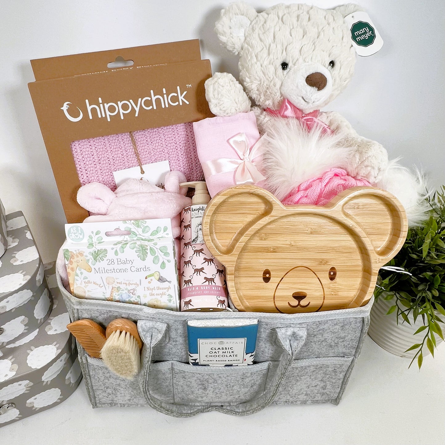 A grey nappy caddy baby hamper gift containing a Mary Meyer large Putty cream baer, a pink baby dressing gown , apink Hippychick cellular baby blankets, a bear bamboo baby plate, a wooden baby hairbrush and comb set a pink pom pom baby hat and various baby toiletries.