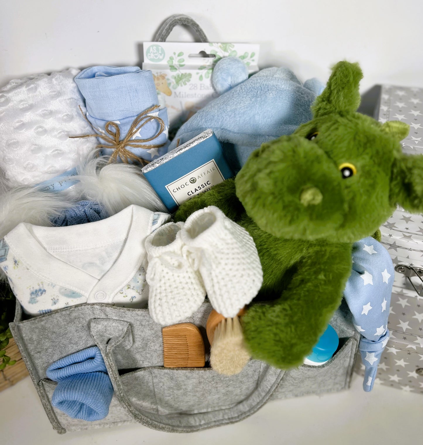 grey nappy caddy containing a green dragon soft baby toy, a blue and white baby sleepsuit, a blue and white baby pompom hat, a soft white baby blankets, a blue baby dressing gown,a ziggle not hat and Ziggle baby bib in blue and white stars.