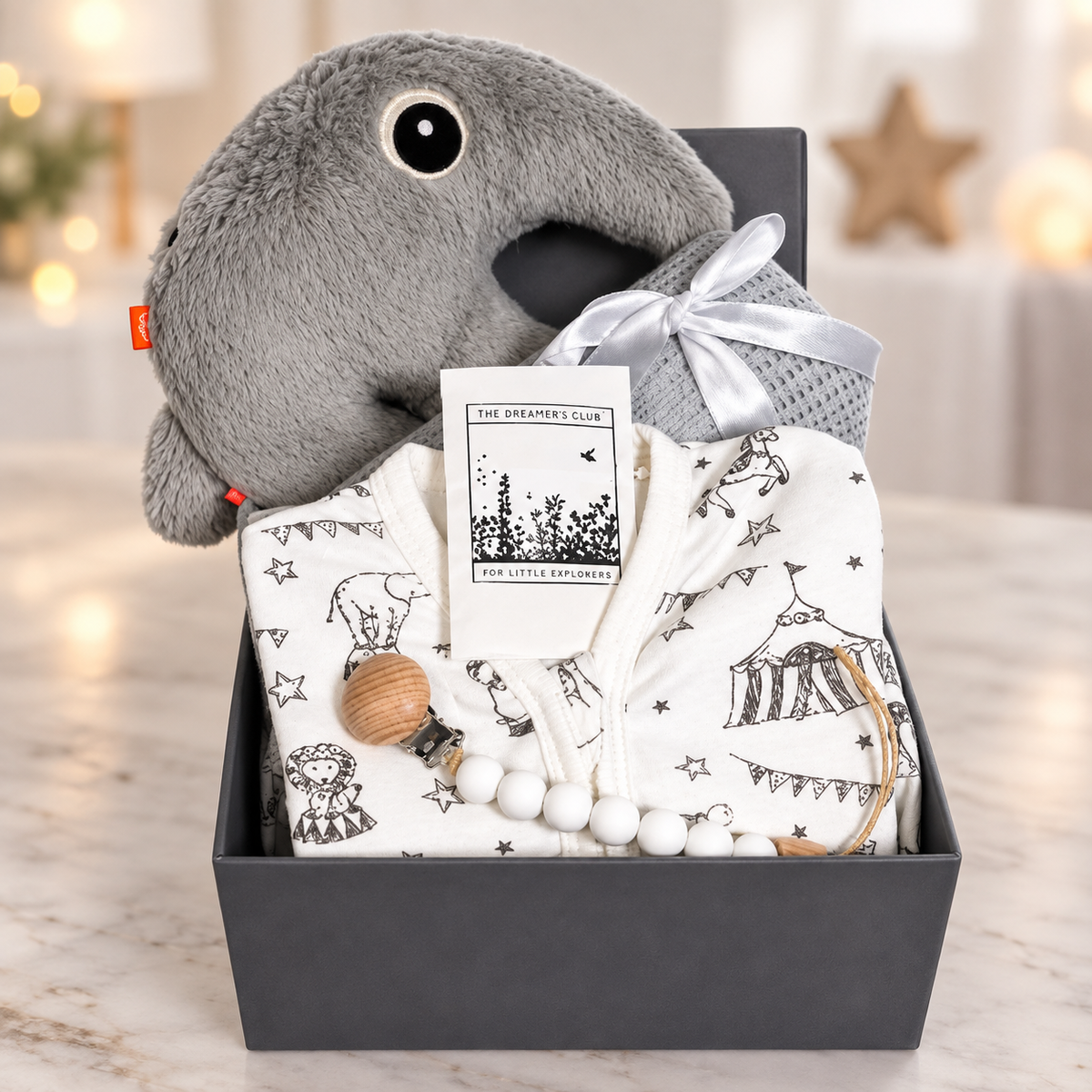 A baby gift hamper in a baby keepsake box featuring a plush ant eater toy, a white printed baby romper with circus motifs, a grey cellular baby blanket tied with a silver ribbon, a dummy clip.