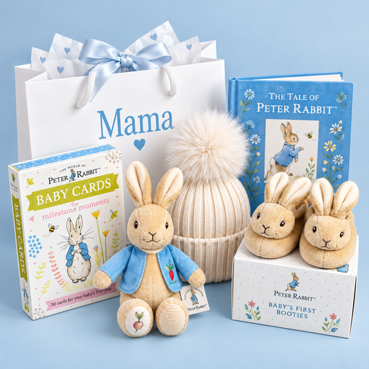 A Peter Rabbit gift set arranged on a light blue background, including a white gift bag with a blue bow and 'Mama' printed on it, a boxed card set, a blue 'The Tale of Peter Rabbit' book, a knitted cream beanie with a large pom-pom, a plush rabbit figure in a blue jacket, and a box of baby booties.