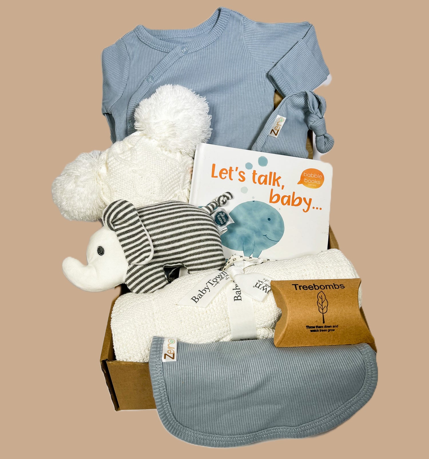 A blue baby boy hamper with a white cotton cellular baby blanket, Mary Meyer baby rattle, blue cotton baby sleepsuit, hat and bib. A white baby pompom hat and a "let talk baby " board book.