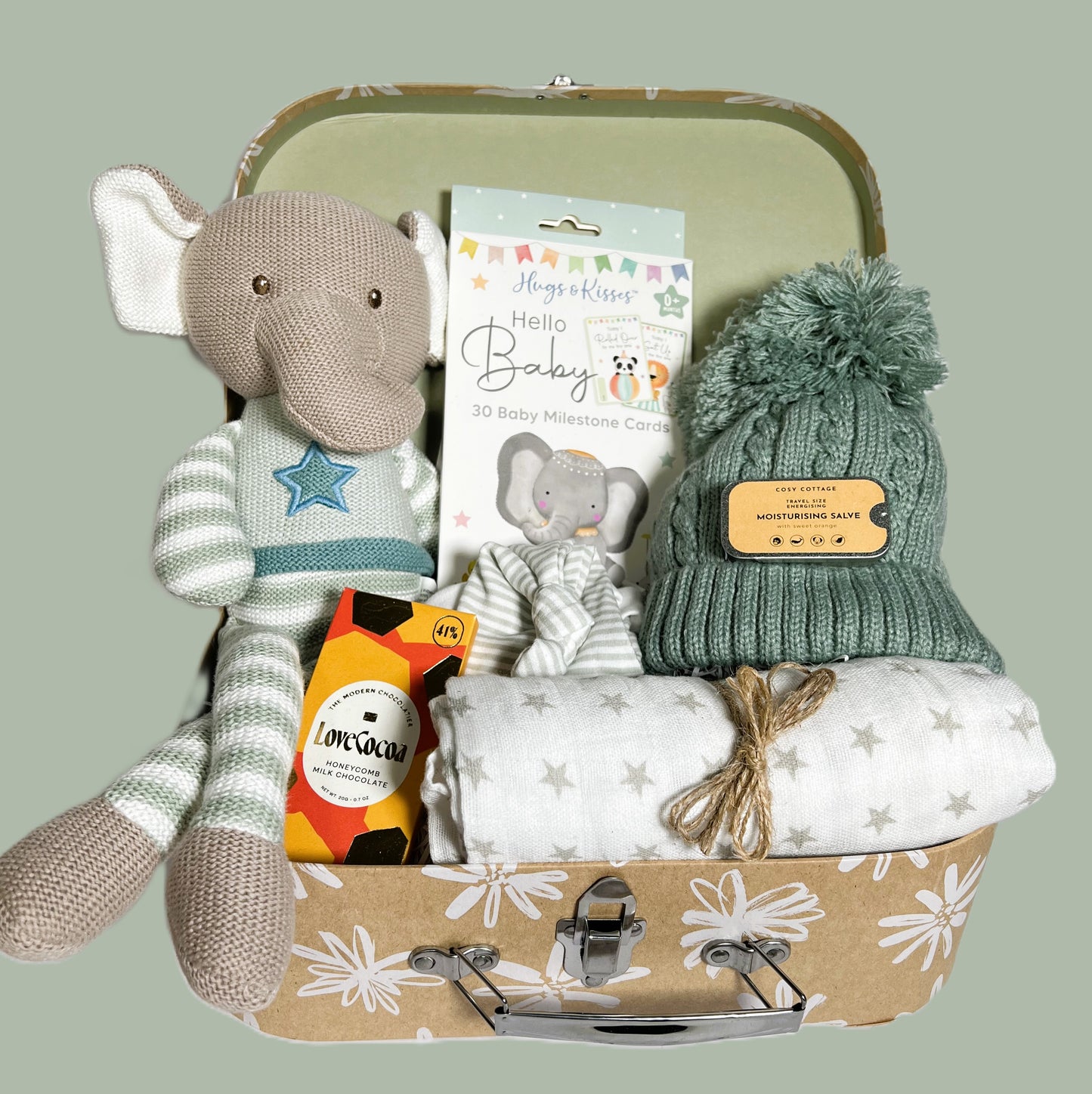 N new baby hamepr with a knitted elephant soft toy, a white muslin square with small star print, a green baby pompom hat, a white and taupe cotton baby knotty hat, a pack of baby milestone cards and some trets for mummy.