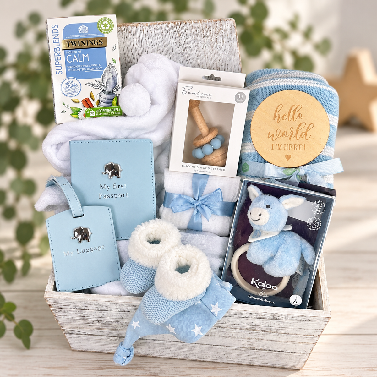 A blue baby boy gift hamper filled with baby items including a blue passport, luggage tag, baby booties, baby blanket, baby robe, teething rings, a plush toy, and  Twinings Calm tea box, arranged in a rustic wooden box  on a light wood surface with blurred greenery in the background.
