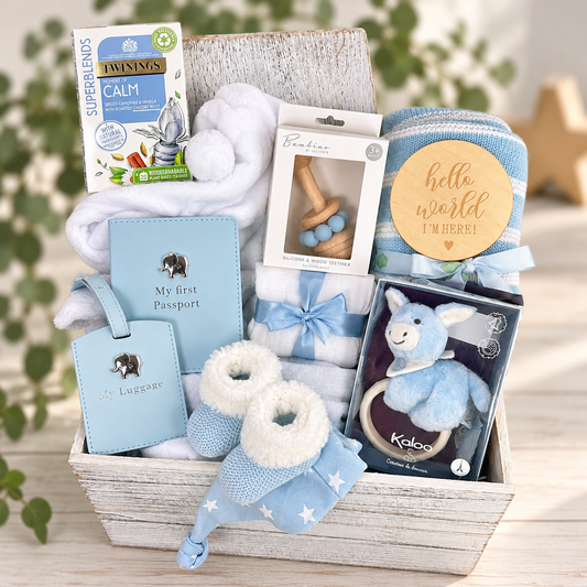 A blue baby boy gift hamper filled with baby items including a blue passport, luggage tag, baby booties, baby blanket, baby robe, teething rings, a plush toy, and  Twinings Calm tea box, arranged in a rustic wooden box  on a light wood surface with blurred greenery in the background.