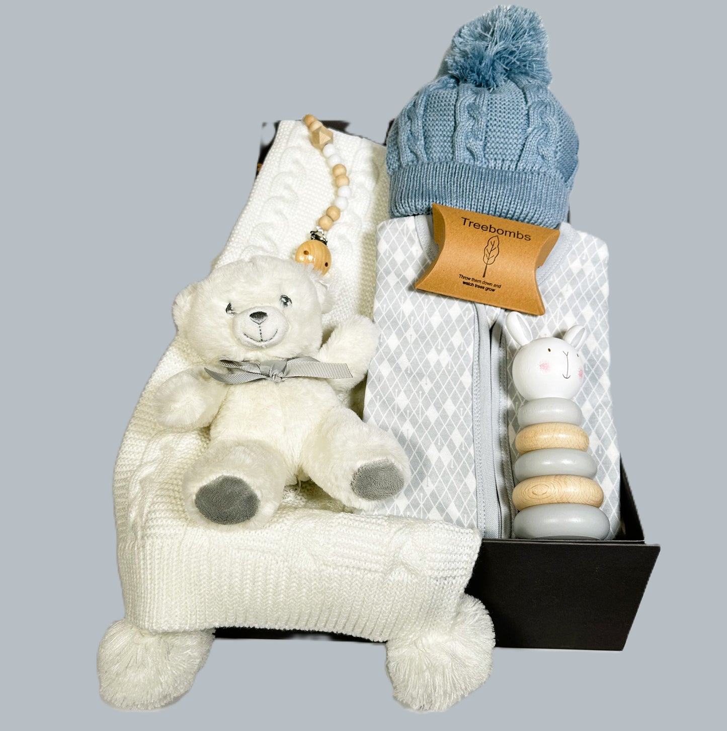 Baby boy hamper in a grey baby keepsake case with a zipped baby sleepsuit, a blue baby pompom hat, a dummy clip, white and grey teddy bear, a white baby blanket and a Bambino stacking baby toy.