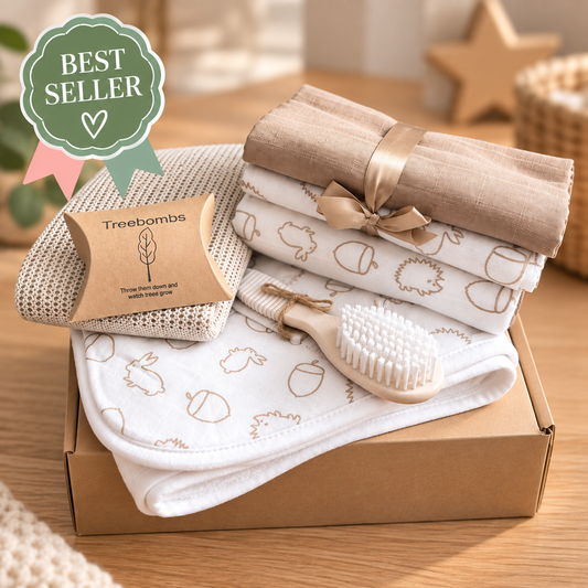 A baby gift set in a brown cardboard box featuring a Treebombs a taupe cotton cellular baby blanket, 3 baby muslin squares tied with a ribbon, a white hooded baby towel with a brown acorn and hedgehog pattern, and a wheatgrass baby brush with white bristles. A green 'BEST SELLER' badge is in the top left on a wooden table background.