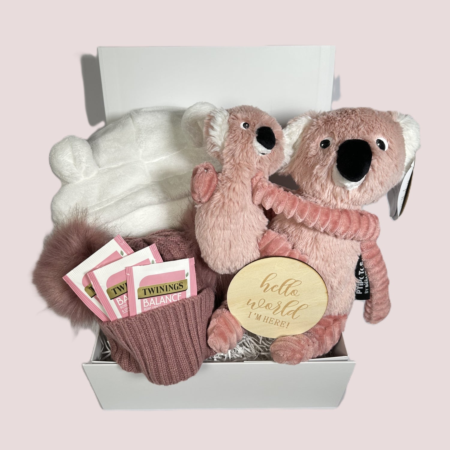 Pink baby girl hamper with a mummy Koala and her baby, a white hooded baby dressing gown, a dusky pink double pompom baby hat, Twinings teabags, a "Hello World" baby photograph sign and a white baby keepsake box.