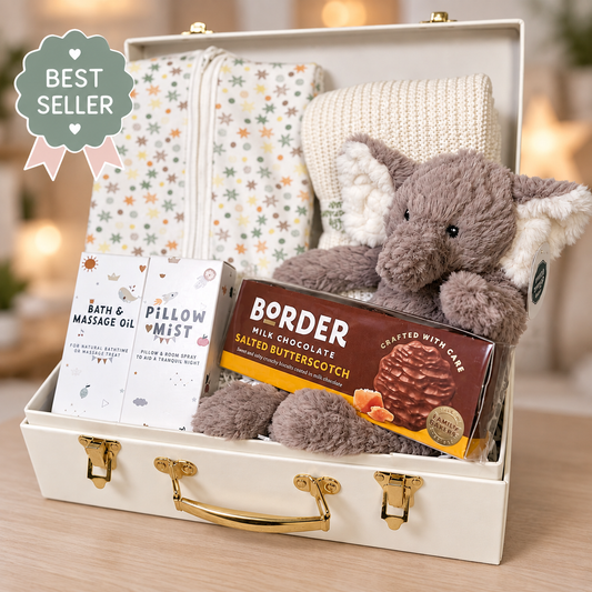 A curated new baby gift box set in a cream-colored suitcase, featuring a plush grey /brown elephant Mary Meyer baby toy, a star print organic cotton baby sleepsuit, a cream cellular baby blanket, a packet of Border milk chocolate biscuits, a bottle of bath and massage oil, and a box of pillow mist. 