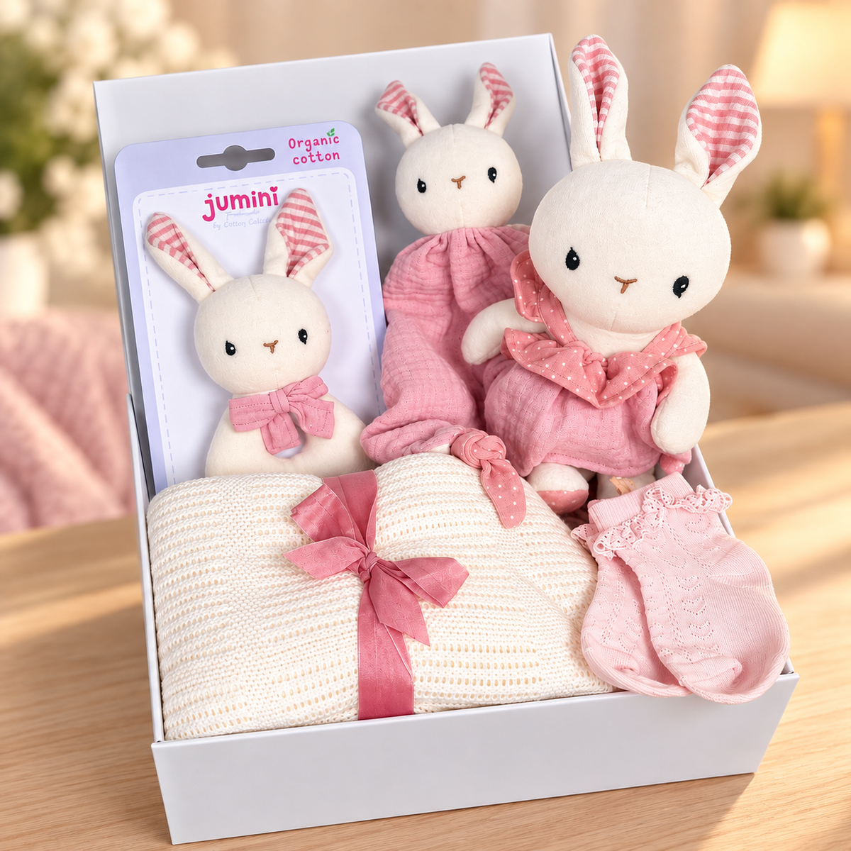 A baby gift set in a white baby keepsake box featuring a plush white bunny doll with pink gingham ears and bow, a matching pink outfit, a cream knit blanket tied with a pink ribbon, and pink booties. A smaller bunny baby rattle in organic cotton.