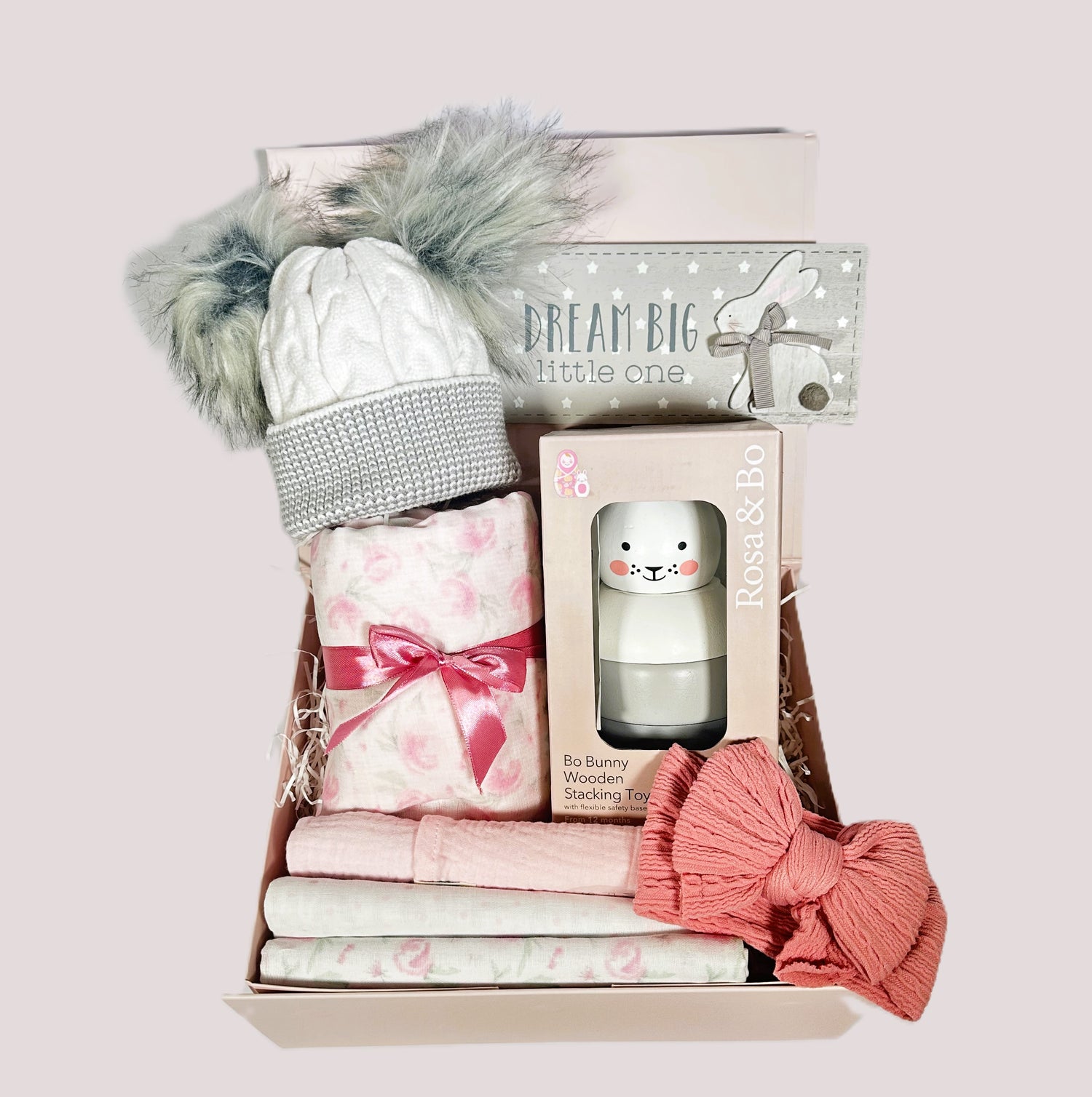 New baby gifts hamper with a pink rose bud baby blanket with cotton waffle to reverse and three matching baby muslins. A Rosa and Bo bunny stacking toy, a pink baby headband, a nursery plaque and a grey and white baby pompom hat.