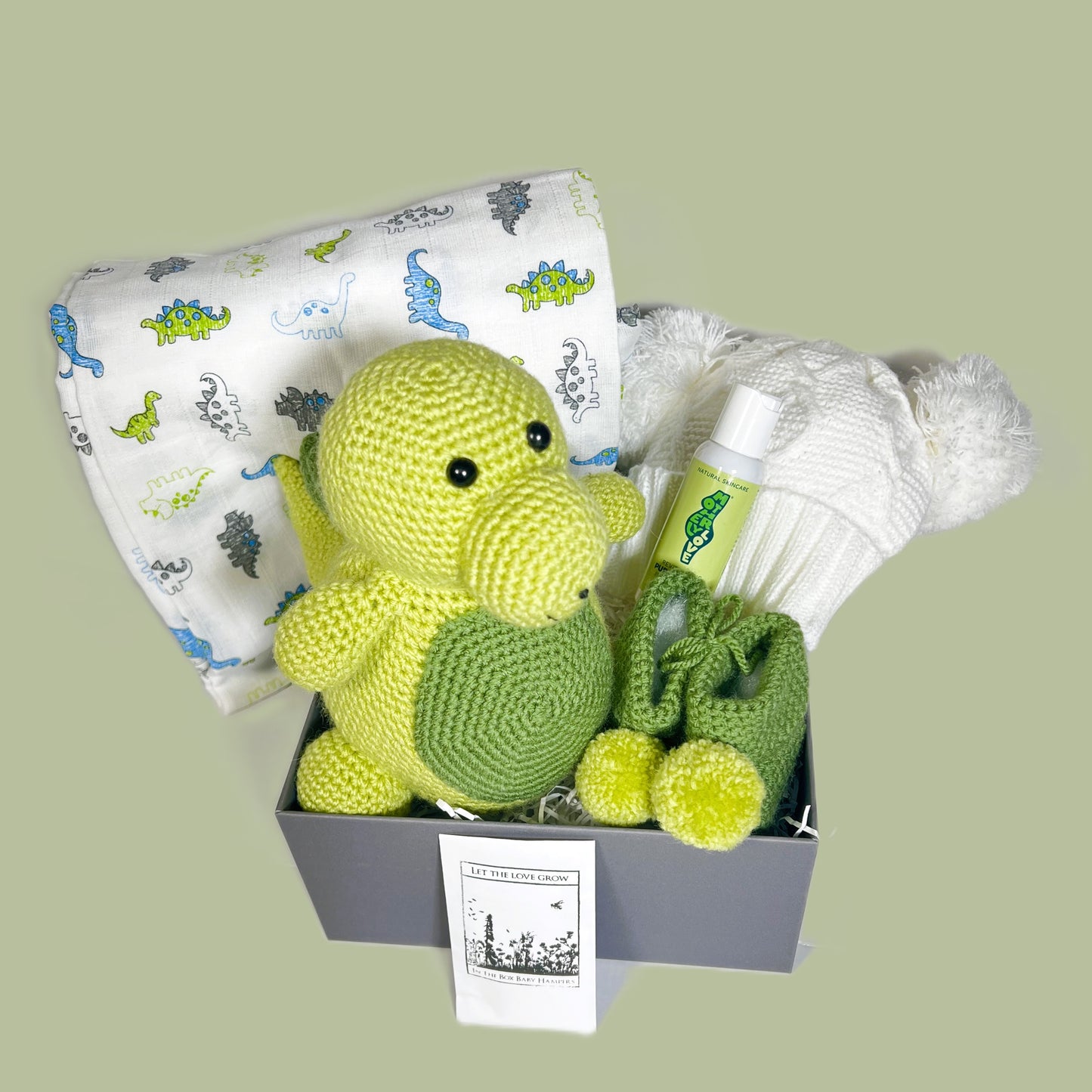 Baby shower gifts. A chrocheted dinosaur with matching pompom baby booties. A white baby muslin with dinosaur print in blue and green. A white double pompom baby hat and a small treat for mummy. All in a grey baby keepsake box with a lid.