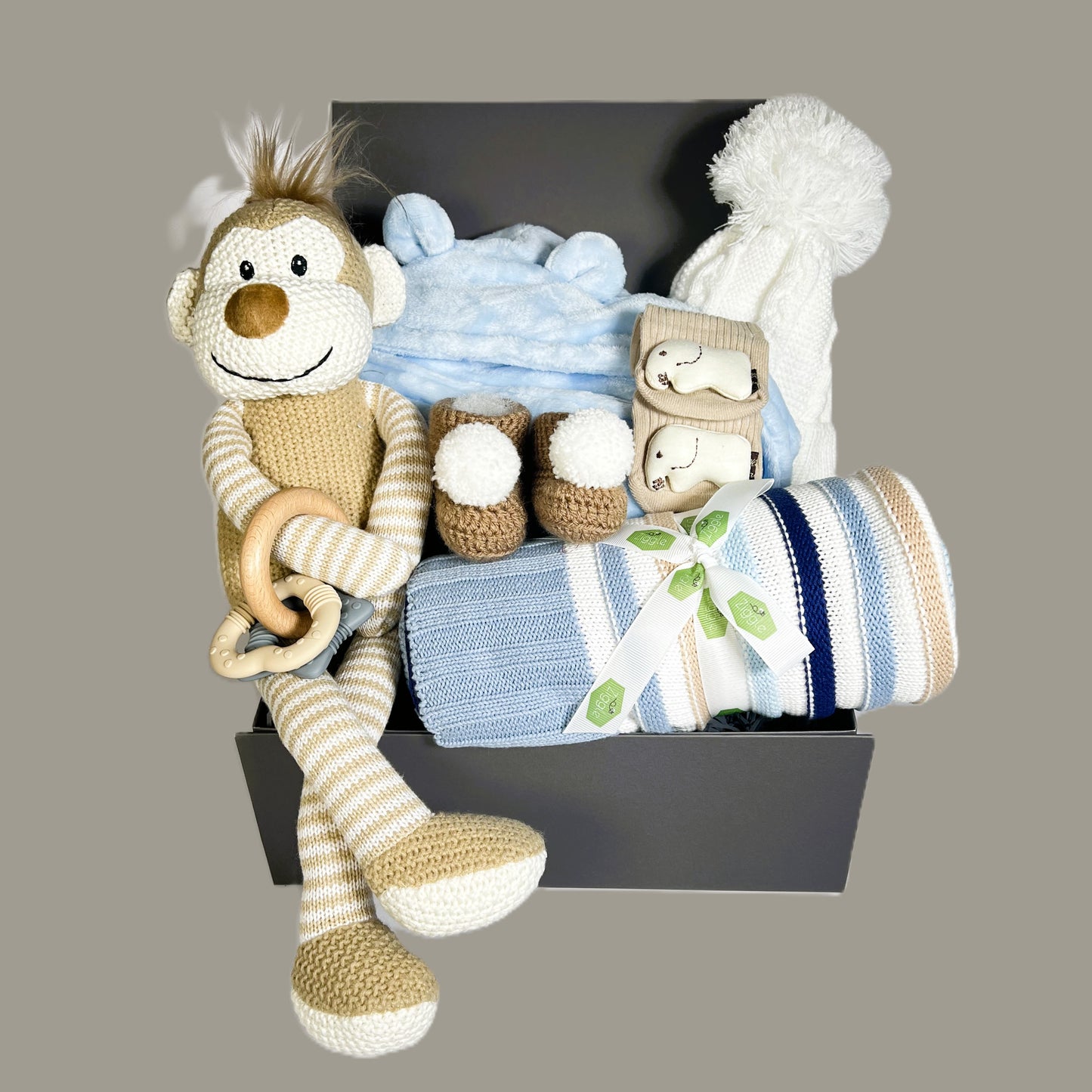 Baby Boy hamper containing a Wilberry knitted monkey baby soft toy, a blue baby dressing gown with a hood, a cotton striped baby blanket. A pair of crocheted baby booties with pompoms, a pair of cotton baby socks with elephant, a baby teething toy and a white baby pompom hat.