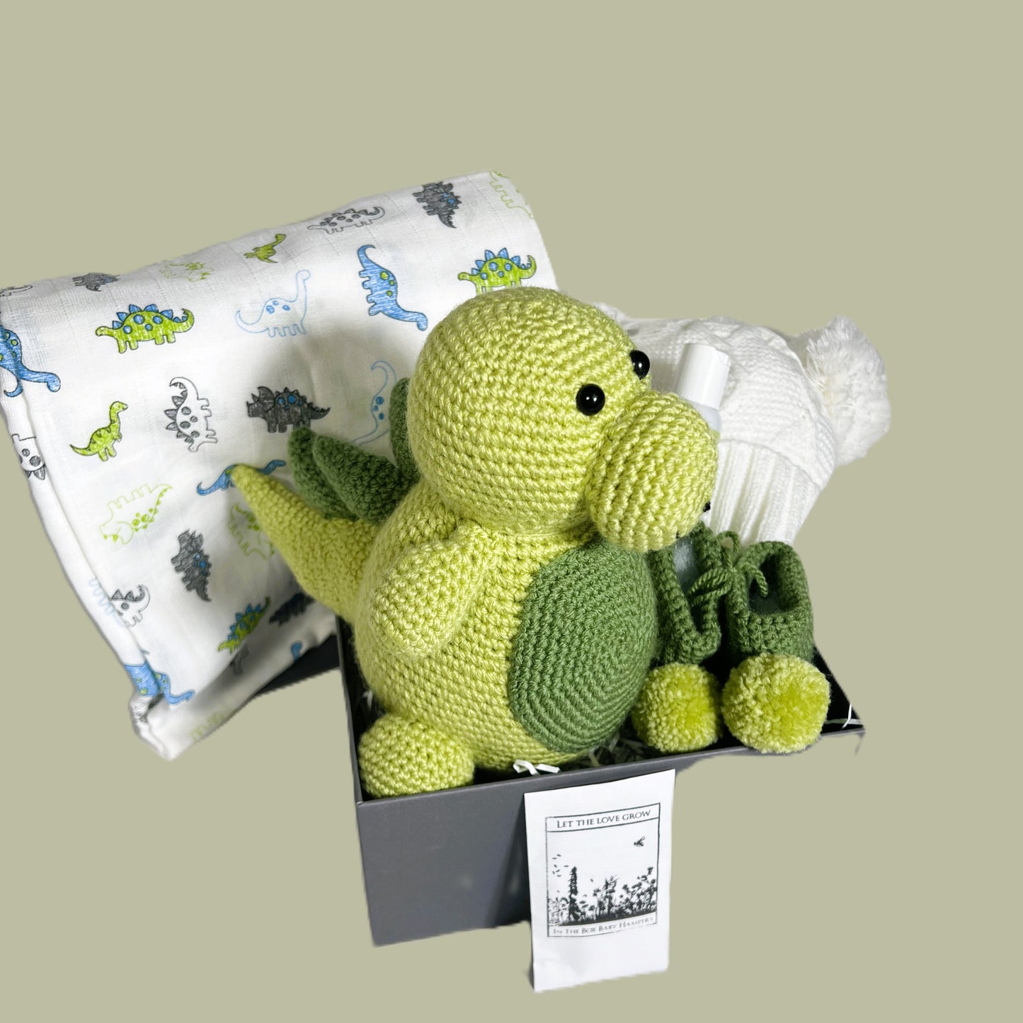 Baby shower gifts. A chrocheted dinosaur with matching pompom baby booties. A white baby muslin with dinosaur print in blue and green. A white double pompom baby hat and a small treat for mummy. All in a grey baby keepsake box with a lid.