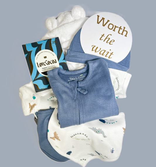 This baby boy hamper comprises a blue baby sleepsuit in organic cotton with a dinosaur print baby bodysuit and matching babby bib and a baby hat, a white baby dressing gown, a bar of Love Cocoa chocolate and a baby keepsake box