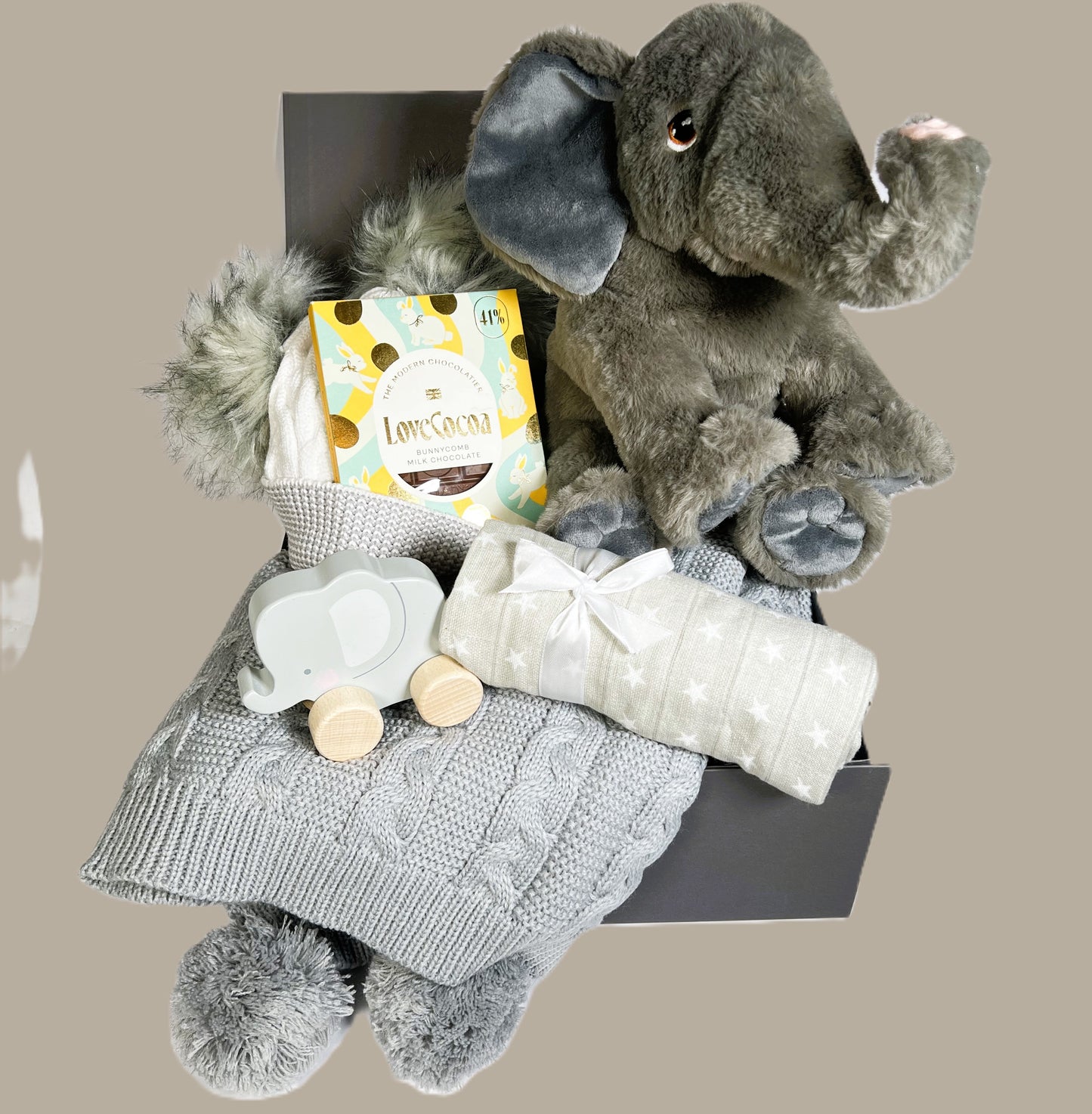 A baby  gift in a baby keepsake box containing an Eco Cuddlies Wilberry elephant soft toy, a grey baby pompom blanket, a push along elephant toy, a bar of Love Cocoa chocolate and a grey and white baby pompom hat.