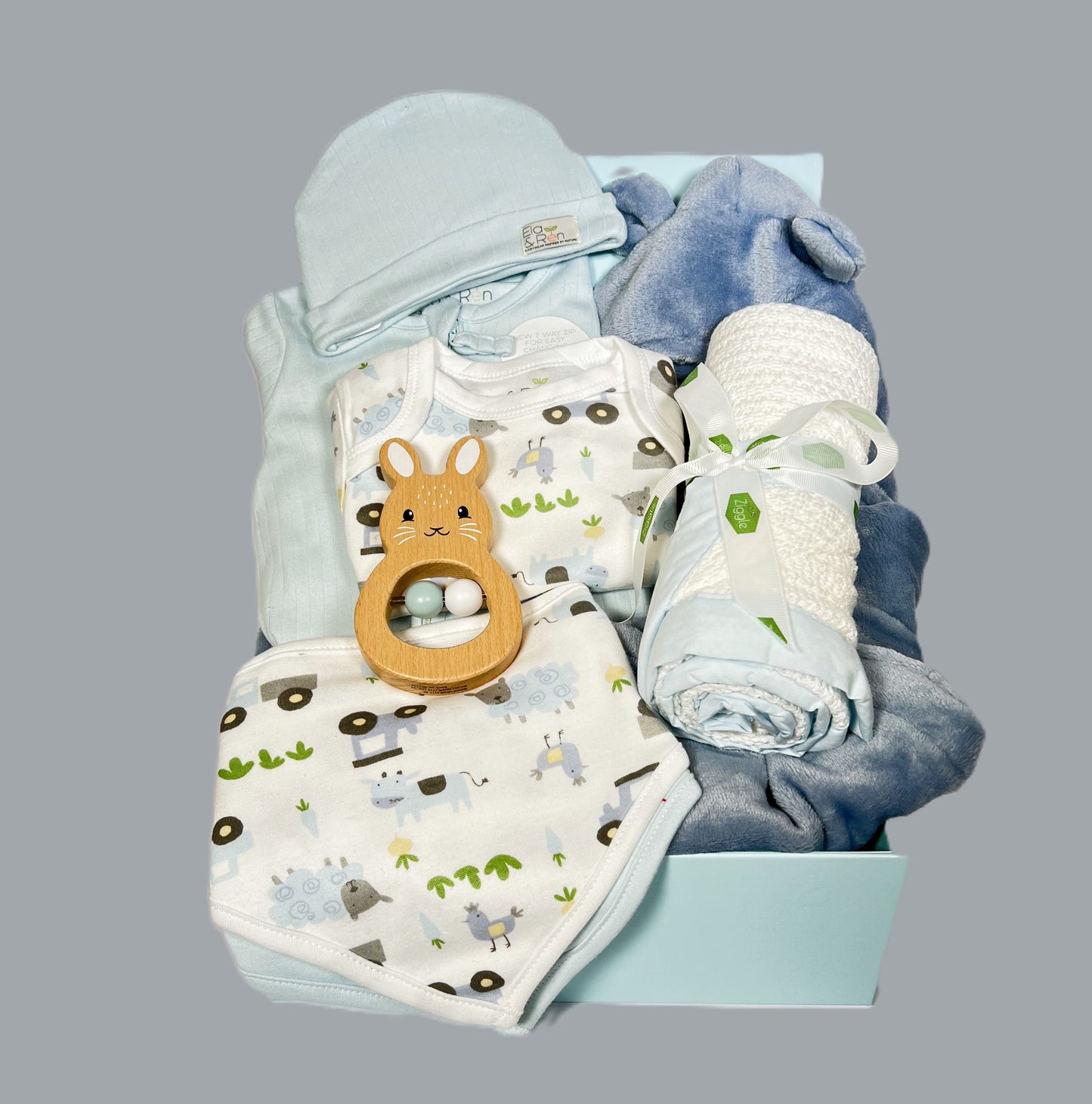 A new baby boy hamper containing Organic cotton baby layette set with double zipped baby sleepsuit, 2 baby dribble bibs, a baby hat and a farmyard print short sleeved baby body suit. A which cotton cellular baby blanket with a blue cotton trim, a wooden bunny baby rattle and a dusky blue baby dressing gown.