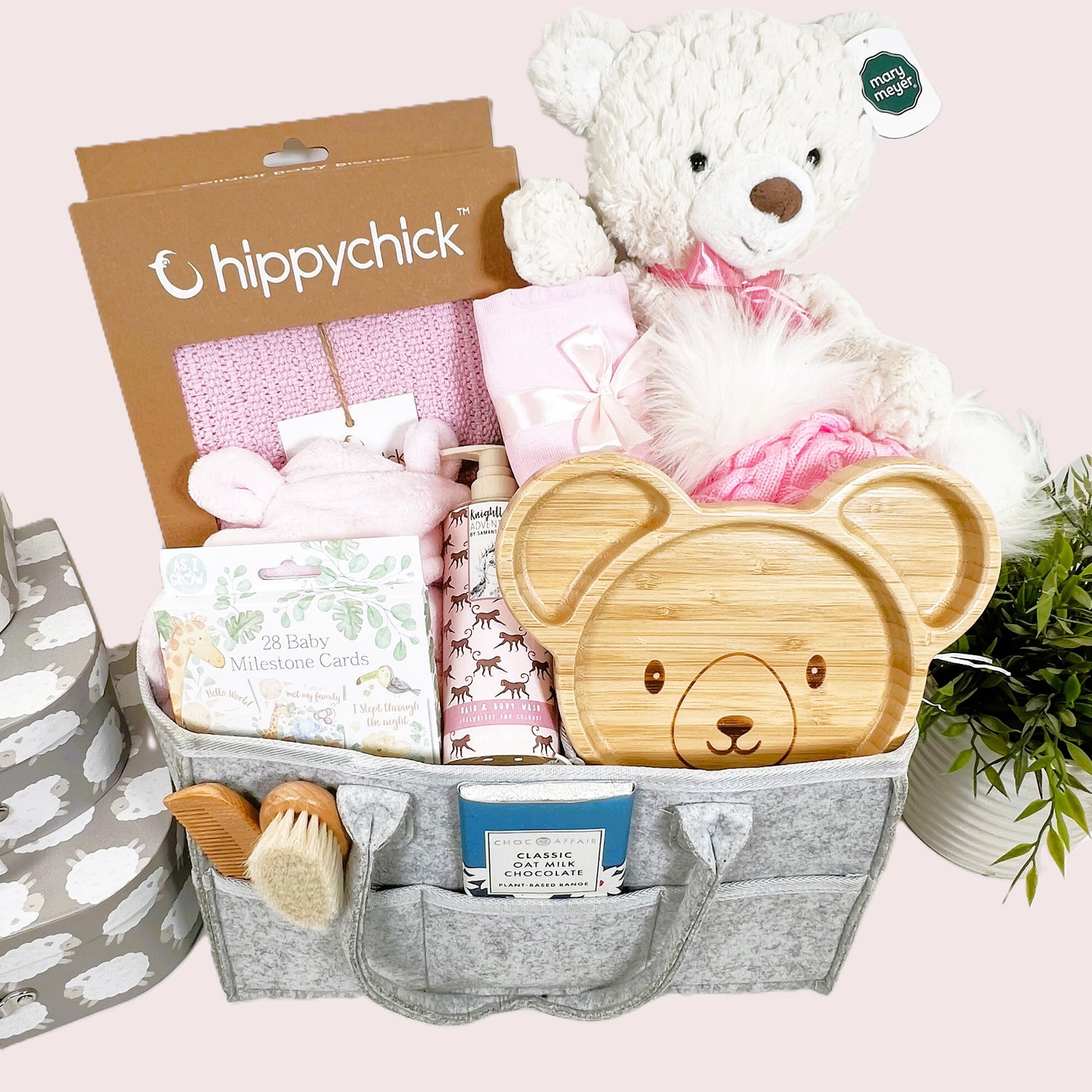 A grey nappy caddy baby hamper gift containing a Mary Meyer large Putty cream baer, a pink baby dressing gown , apink Hippychick cellular baby blankets, a bear bamboo baby plate, a wooden baby hairbrush and comb set a pink pom pom baby hat and various baby toiletries.