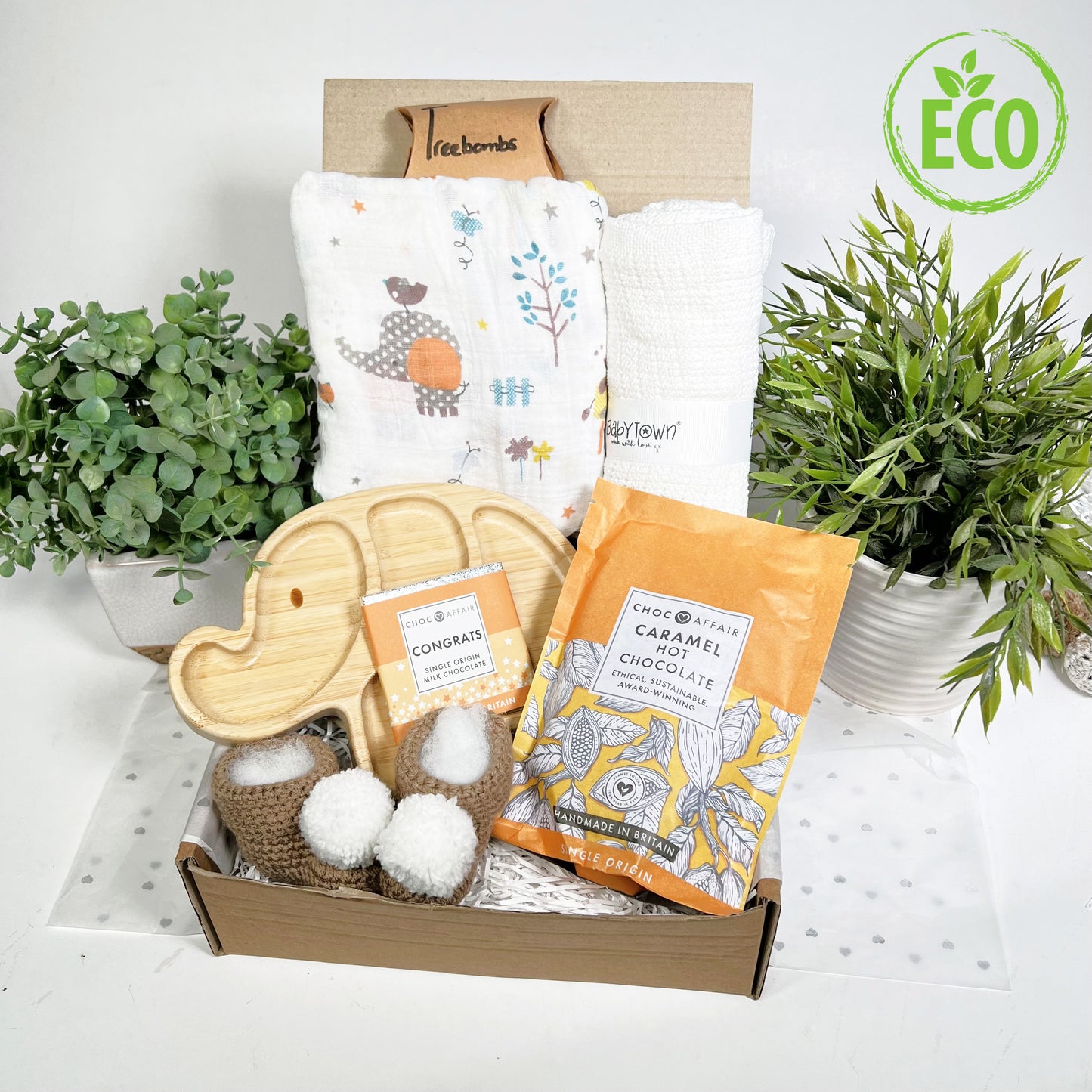 Eco Friendly neutral baby shower gift hamper box containing a bamboo elephant shaped baby plate with 3 internal sections, a large muslin square with a neutral elephant and jungle print, a white cotton cellular baby blanket, a pouch of caramel hot chocolate and a chocolate var with the text "Congrats", a pair of crocheted baby booties with pompoms and a free packet of 3 Treebombs.