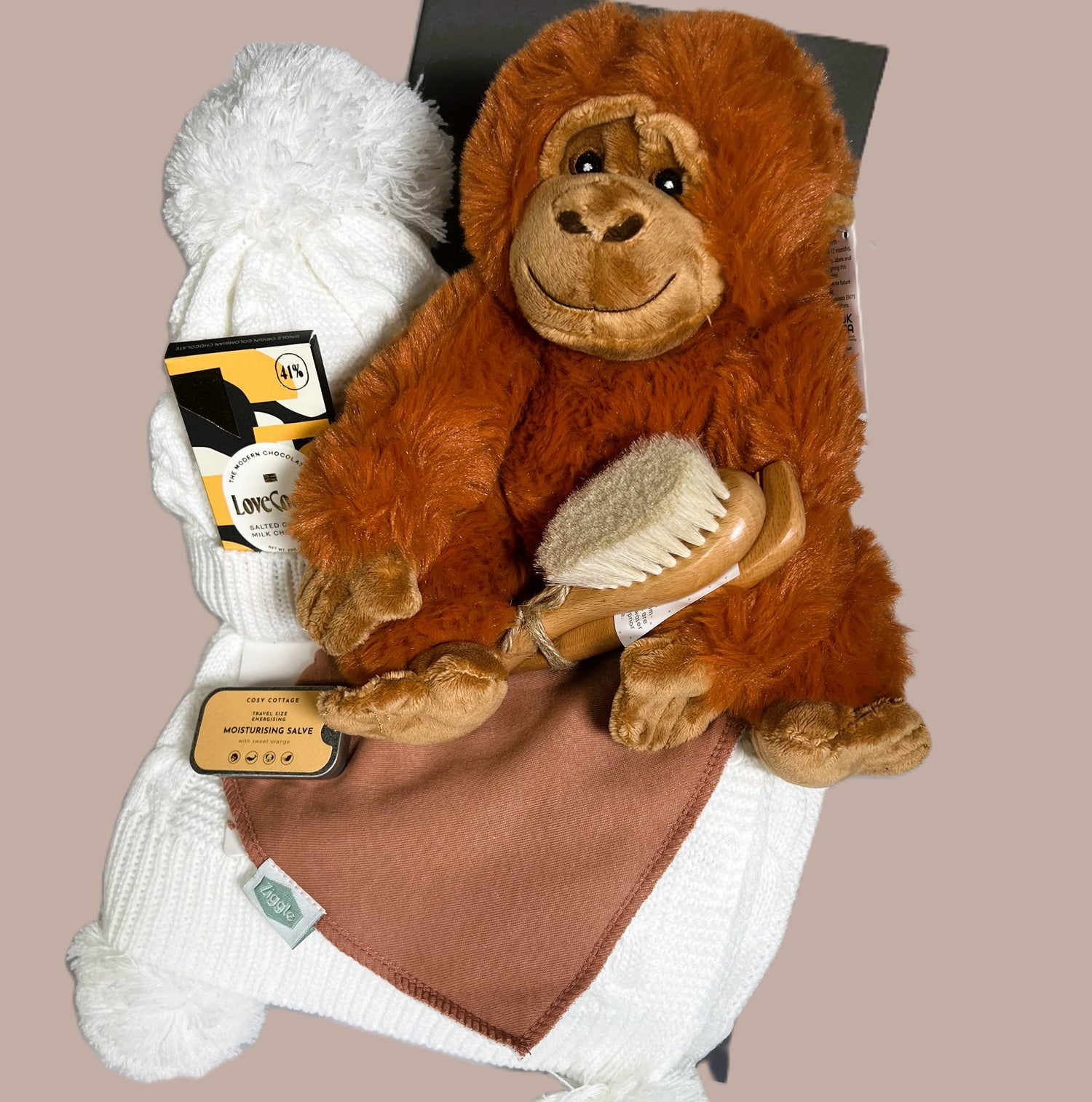 A baby shower gift in a baby keepsake box containing an orangutang soft baby toy, a white cable knit baby blanket and matching pompom baby hat. A wooden baby brush and comb set, a pot of Cost Cottage moisturising slave, a bar of Love Cocoa chocolate and a baby dribble bib.