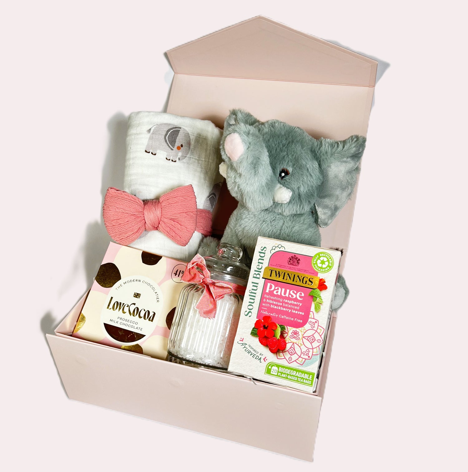 Pink baby girl hamper containing a white large swaddle blanket with an elephant print, a Wilberry eco cuddlie Ela elephant soft baby toy, a bar of Love Cocoa  prosecco flavoured chocolate, a box of Twinings Pause teabags, a baby head band and a jar of coconut bath salts in a pink magnetic baby keepsake box/