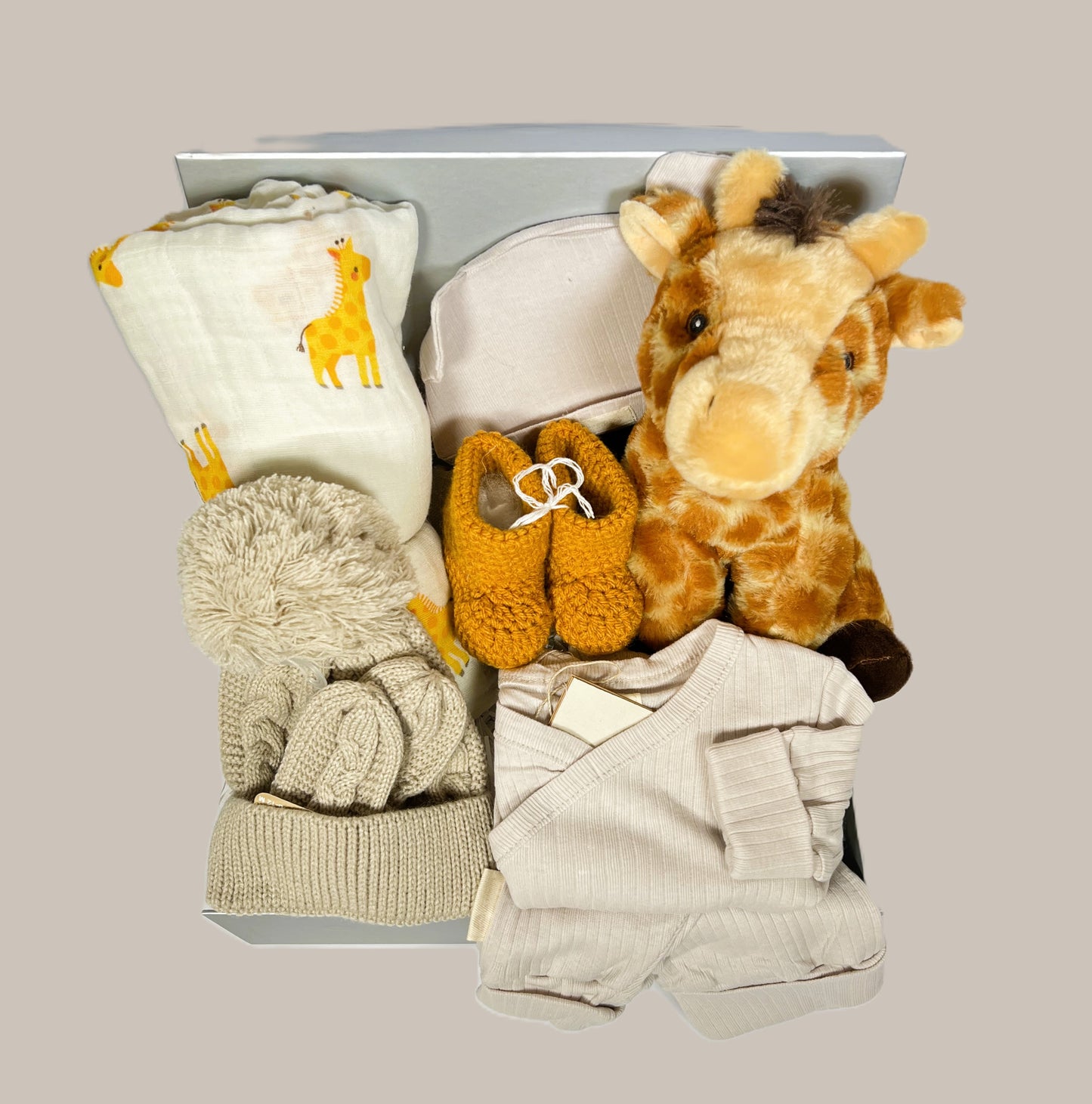 New baby hamper with an organic cotton baby outfit in taupe, a biscuits coloured baby pompom hat and matching mittens. A pair of mustard crocheted baby booties. A large giraffe print muslin  and Wilberry eco cuddlies giraffe soft baby toy.