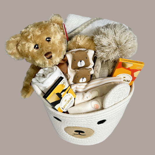 A cream rope storage caddy with a Keeleco teddy, a hooded baby towel, a baby pompom hat, a set of cotton baby clothing, 2bars of Love Cocoa minis, a pair of baby sock and a muslin square.