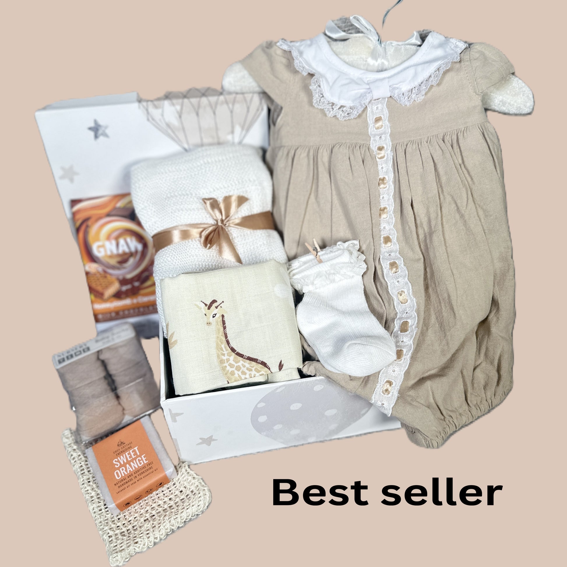 A baby gift hamper arranged on a beige background, including a beige and white baby outfit, a folded white bamboo baby blanket, giraffe-print muslin square, baby socks, and new mummy treats. 