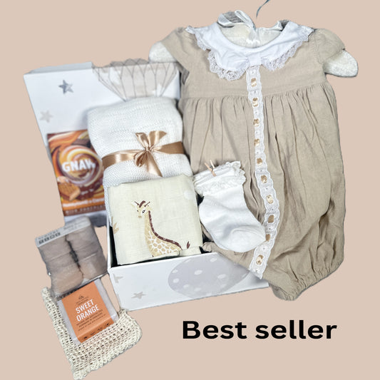A baby gift hamper arranged on a beige background, including a beige and white baby outfit, a folded white bamboo baby blanket, giraffe-print muslin square, baby socks, and new mummy treats. 