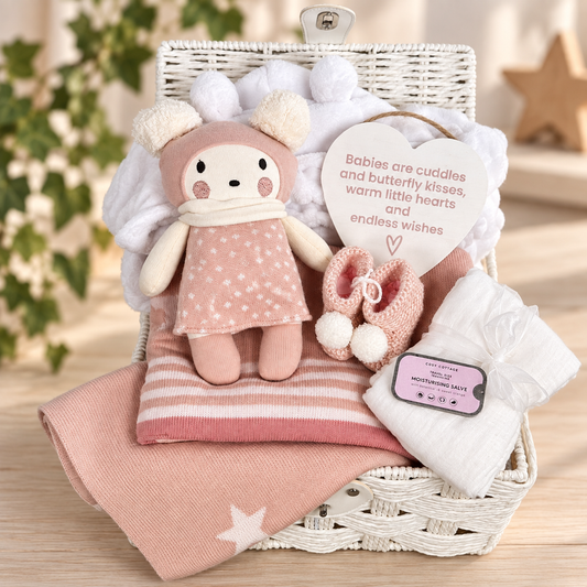 A baby gift basket set in a white paper rope hamper , containing a pink cotton baby toy with cream pom-poms, a pink and white striped  cotton baby blanket, a folded white baby muslin, crocheted  pink  and white baby booties. A heart-shaped nursery plaque and a tin of salve, set against a softly blurred background of greenery and a wooden star.