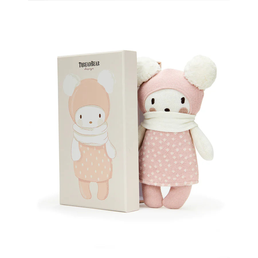 Bella soft knitted baby toy with pink outfit and white hat next to its packaging on a white background