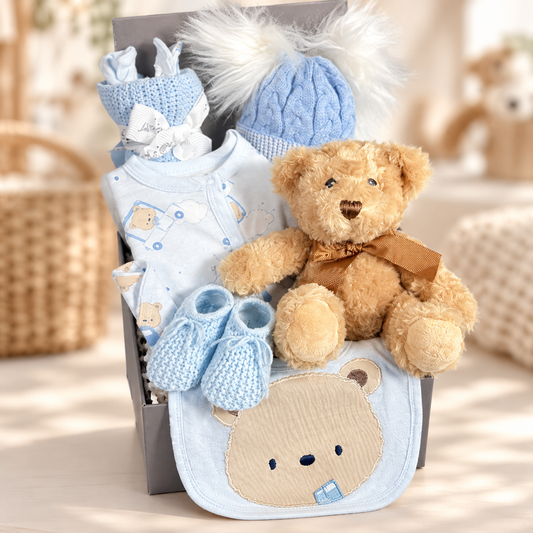 A baby hamper for a boy in a square grey magnetic baby keepsake box containing a blue bear print cotton layette set including a romper suit, baby hat, bodysuit and scratch mitts. A double pompom blue baby hat, a white muslin square a blue cotton cellular baby blanket and a Keeleco Dougie bear with a pair of blue baby bootees.