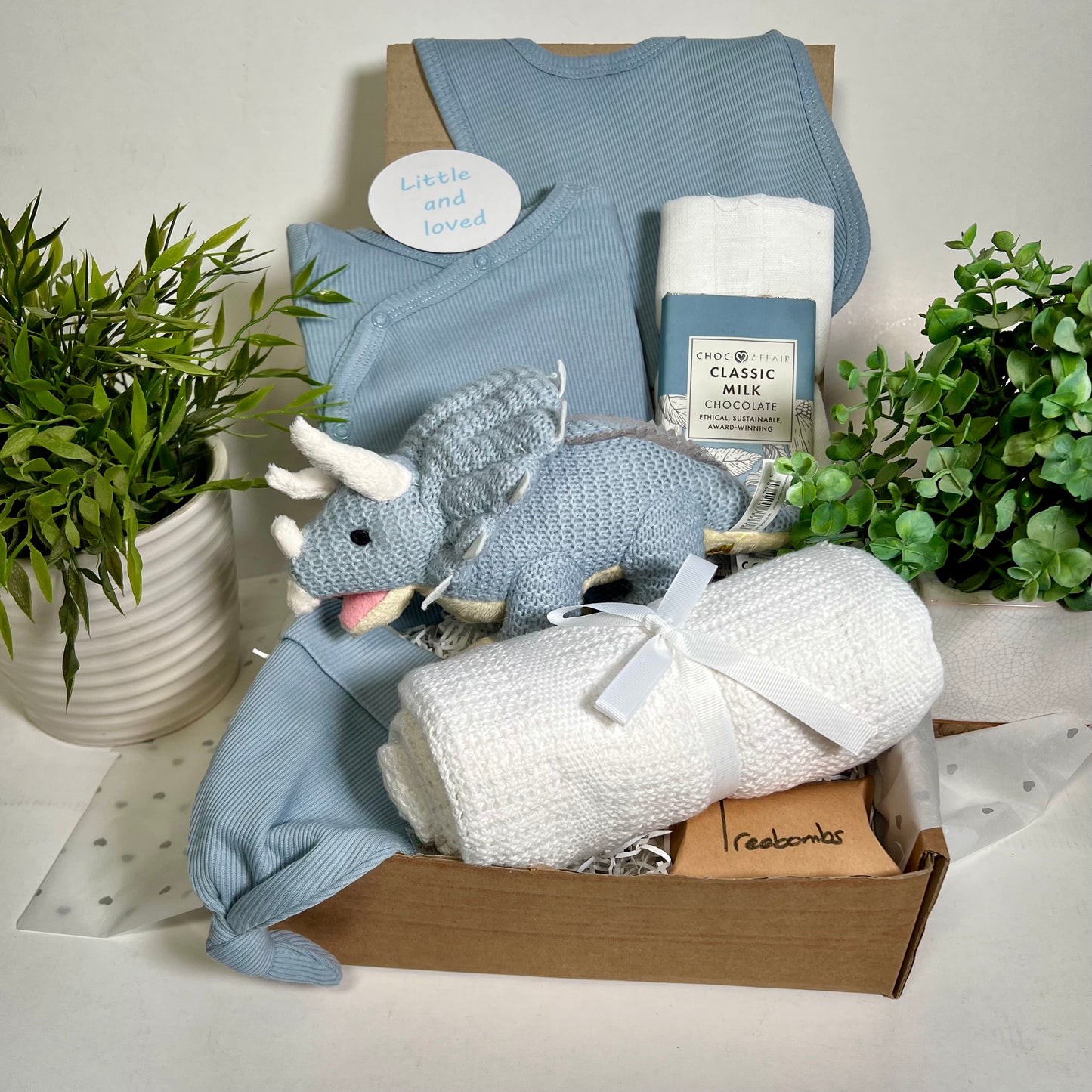 New baby boy gift comprising a blue knitted triceratops dinosaur soft toy, a white  cotton cellular baby blanket, a white muslin square, a blue coton baby clothing set incluing a sleepsuit, dribble bib and bay knot hat,  a Bar of Choc Affair milk chocolate a smapp reveresible baby photography disc and a pack of Treebombs.