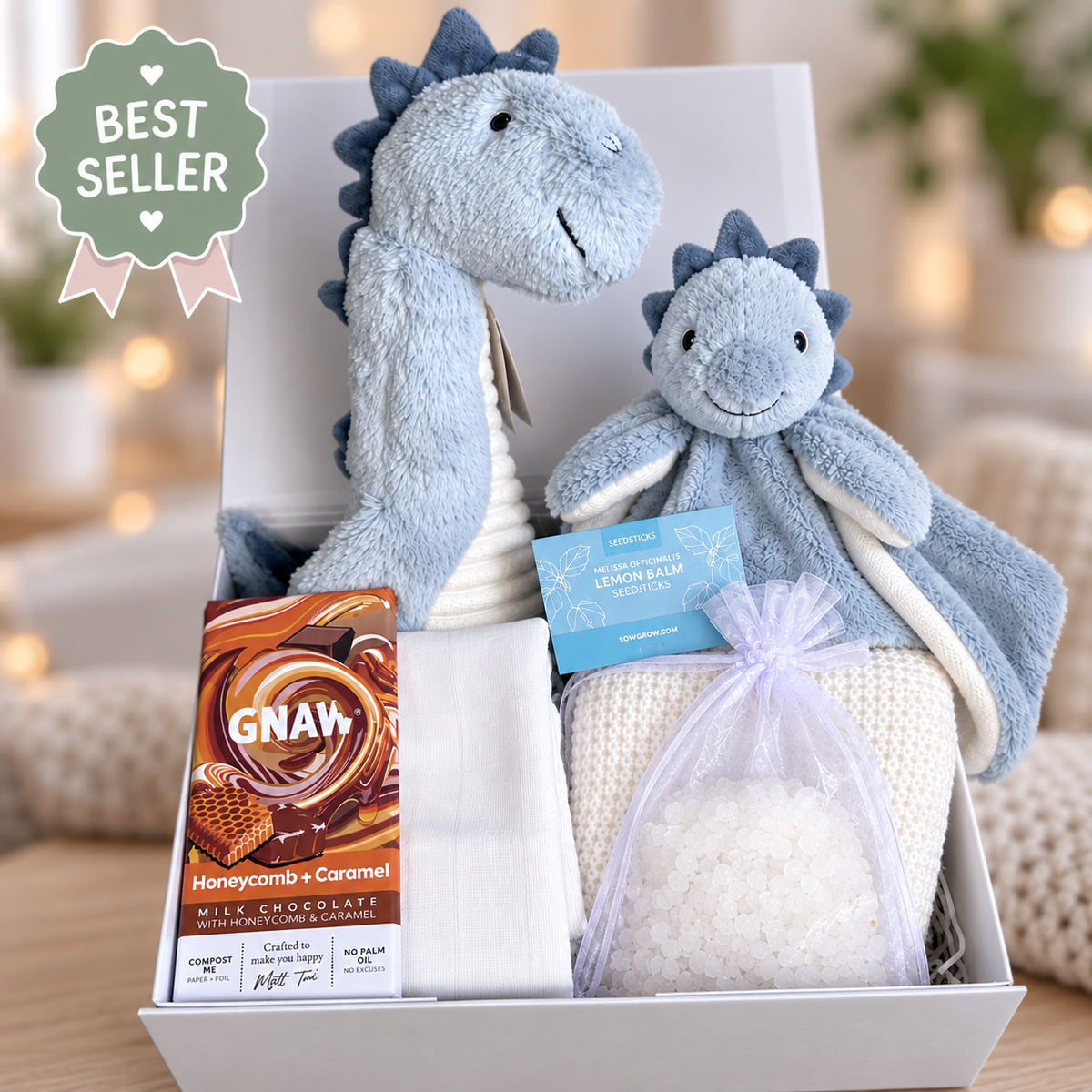 A curated new baby gift hamper featuring a plush Dino blue dragon toy, a matching Dino dragon lovey, a bar of GNAW Honeycomb + Caramel milk chocolate, a white cotton cellular baby blanket and white muslin, and a bag of coconut bath salts, all in a white baby keepsake box.