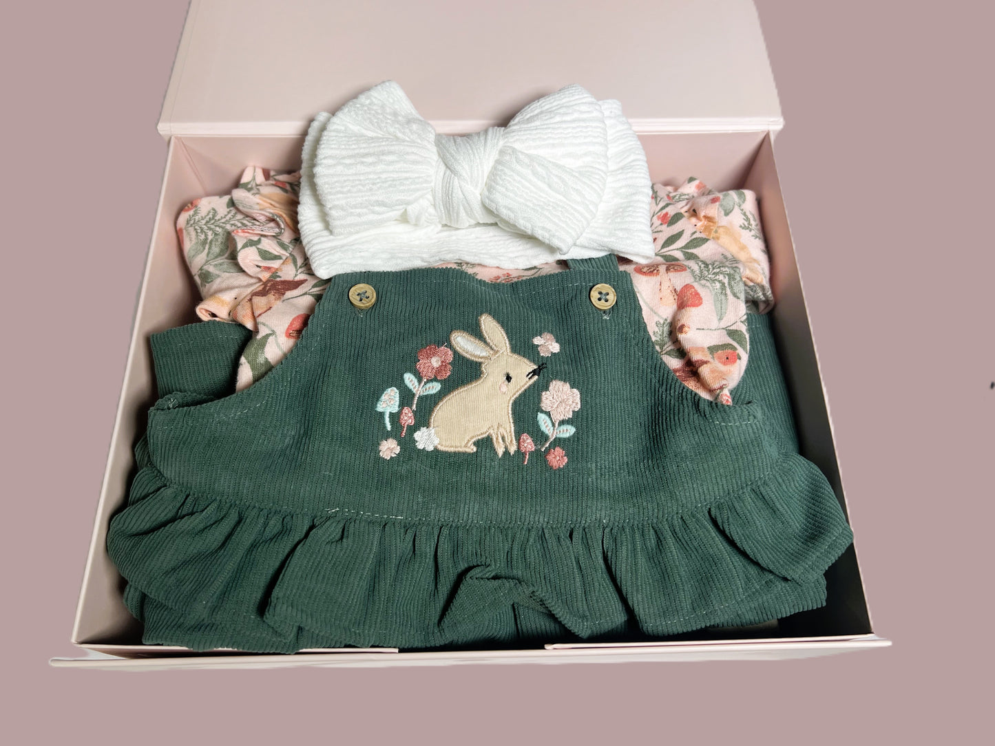 A new baby girl hamper with a corduroy pinafore dress and body suit with long sleeves and matching peach coloured baby tights. A pair of crocheted baby booties with pom poms. A white baby bubble blanket a, wood nursery ornament and a floral print baby muslin square.