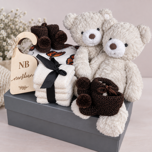 A baby gift hamper arranged on a grey box, featuring two cream plush teddy bears, 2 pairs of dark brown and copper crocheted booties, folded cream reusable baby wipes  tied with a black ribbon, and a wooden baby wardrobe dividers.