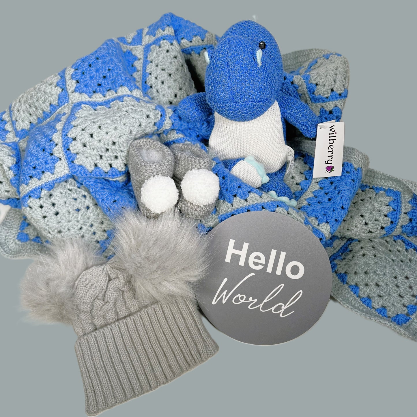 Grey and blue granny square baby boy crocheted baby blanket with a pair of grey baby booties with pompoms, a grey baby pompom hat, a "Hello World" baby photography plaque.