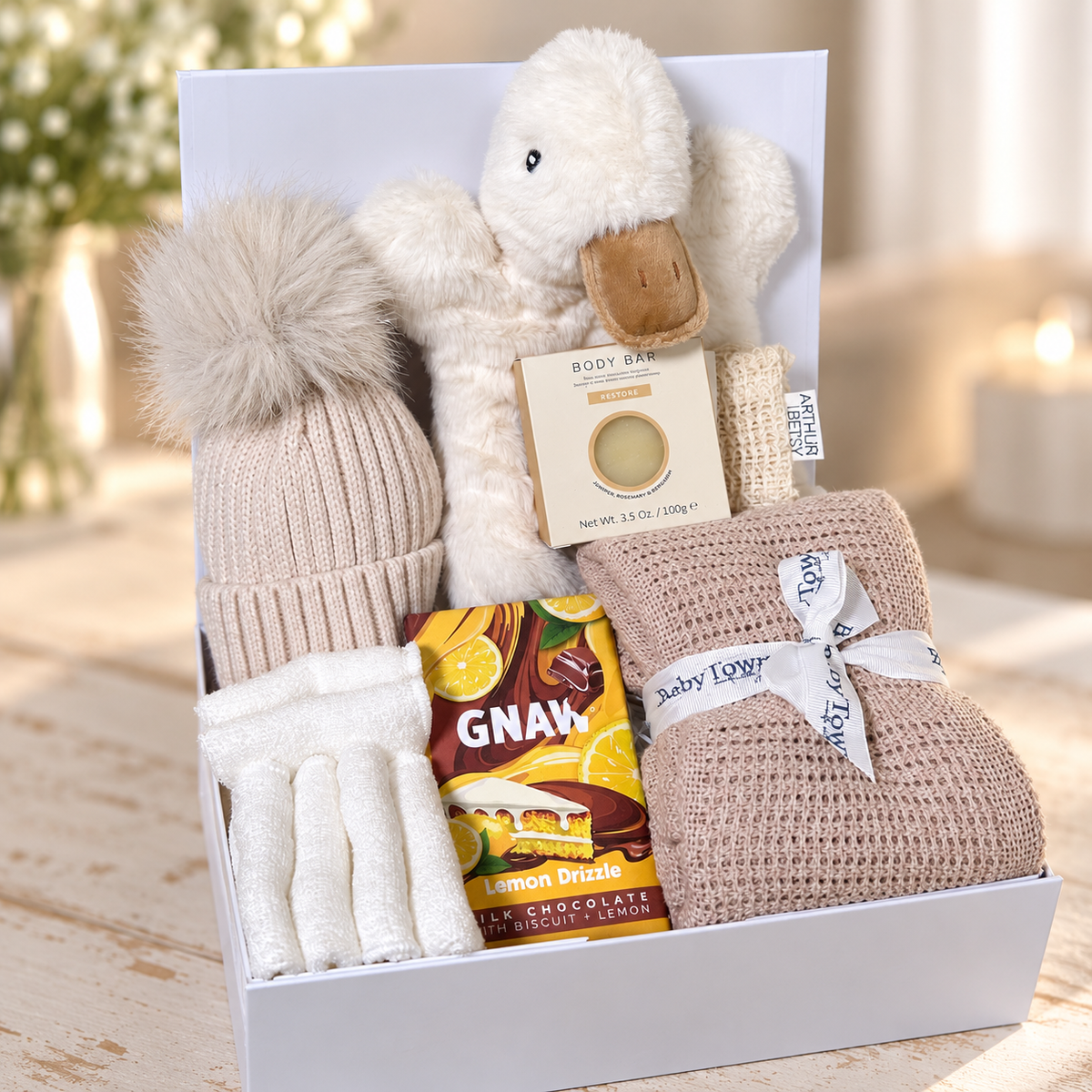 A baby gift hamper in a white baby keepsake box containing a cream plush Goose baby comforter/ hand puppet toy, a beige knit pom-pom hat, white reusable baby bamboo wipes, a brown and yellow GNAW chocolate bar, a folded beige cotton cellular baby  blanket tied with a white ribbon, and a small soap bar and sisal soap bag arranged on a light wooden surface.